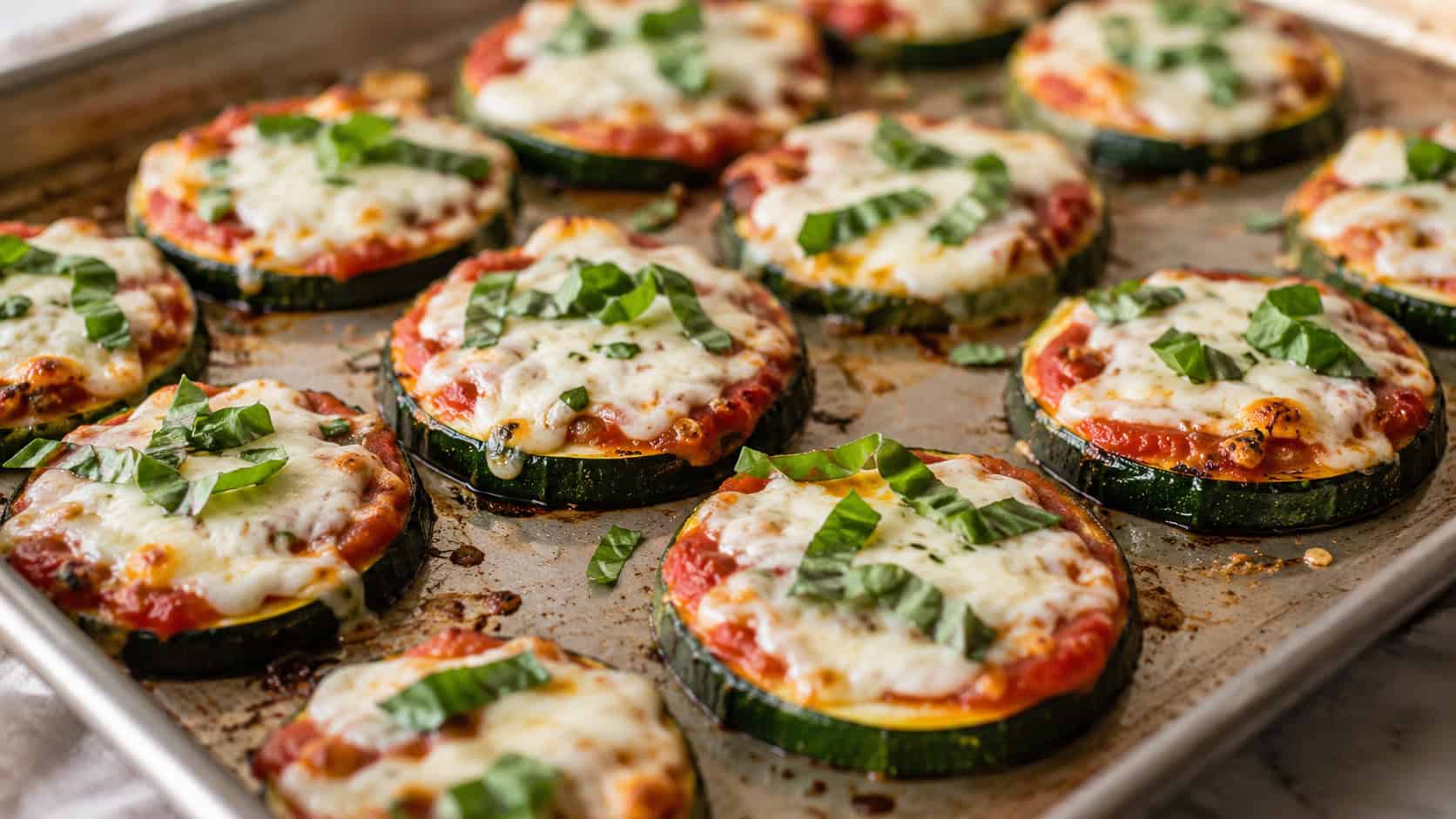 mini zucchini caprese pizzas arranged on a baking sheet in a wide horizontal shot, melted cheese, red marinara, fresh basil leaves, warm natural light, shallow depth of field, landscape orientation