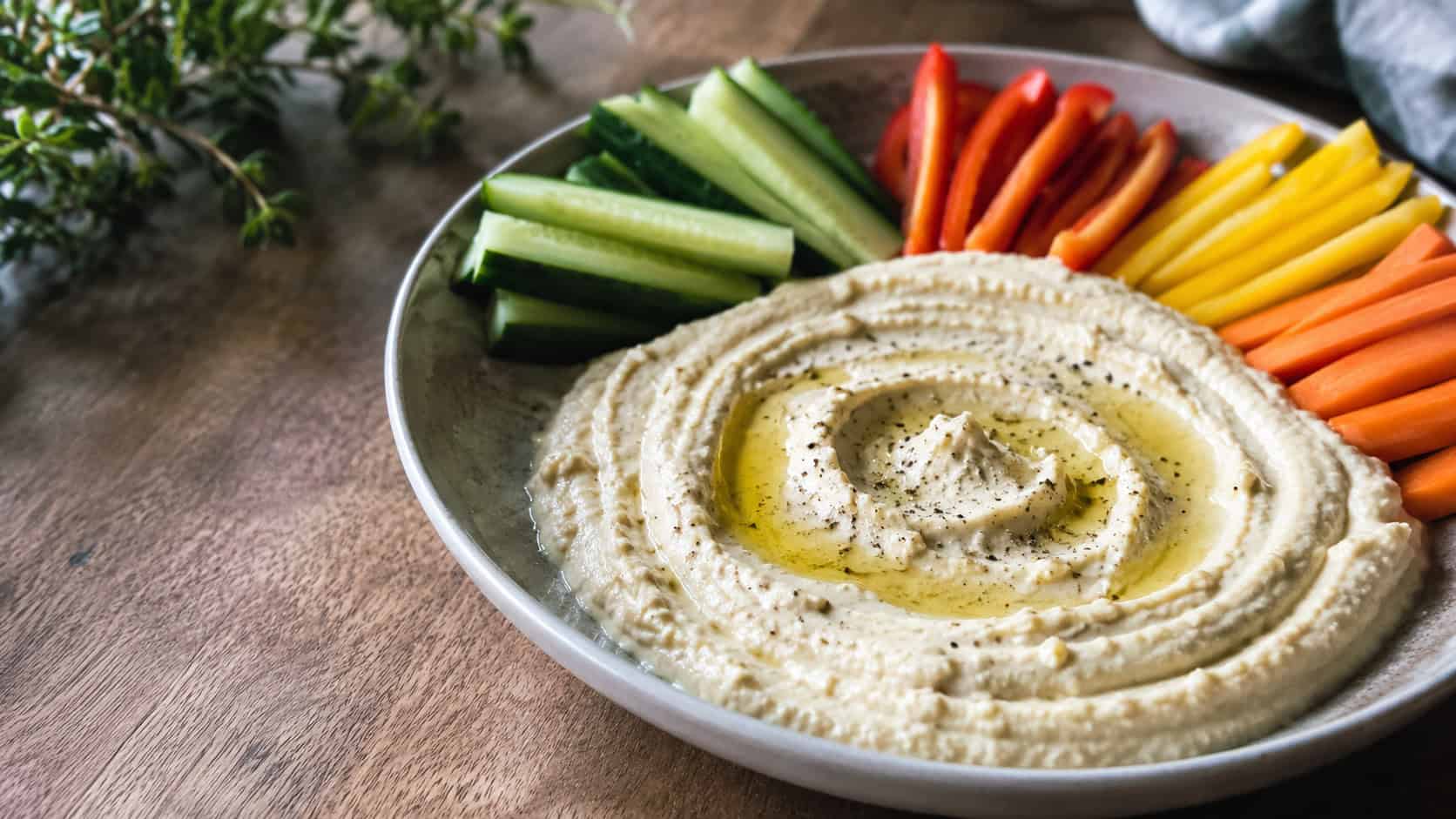 creamy cauliflower hummus swirled in a shallow bowl in a wide horizontal shot, drizzle of olive oil, cucumber and pepper sticks fanned alongside, soft natural light, shallow depth of field, landscape orientation