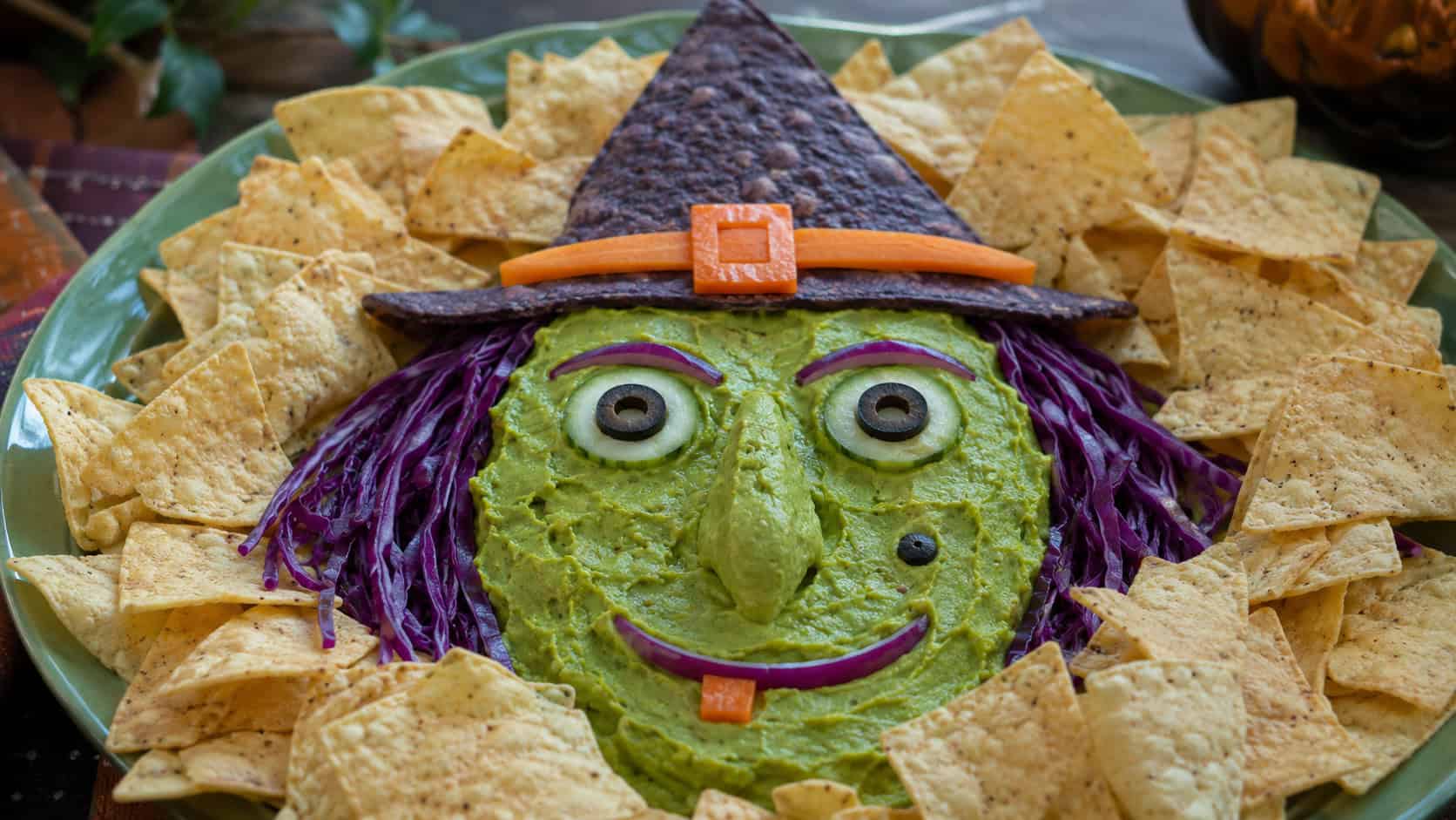 guacamole spread into a witch's face on a platter with a tortilla-triangle hat and veggie features in a wide horizontal shot, on a green platter surrounded by tortilla chips, natural light, shallow depth of field, landscape orientation