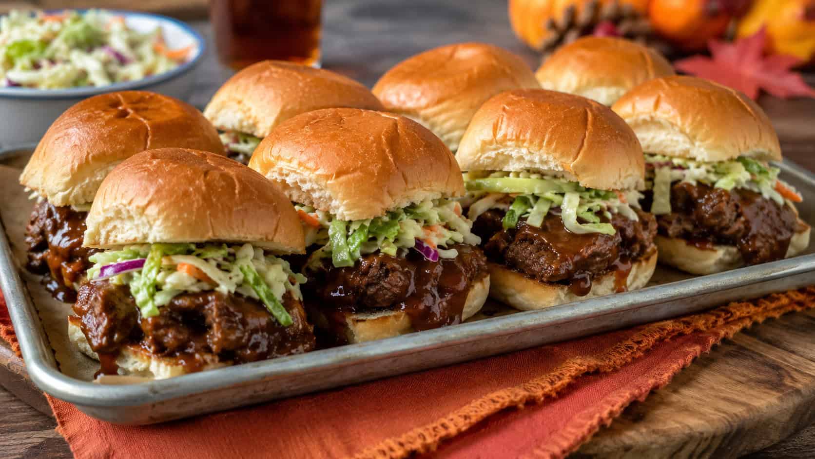 a tray of saucy beef sliders with bright green slaw spilling out of soft buns in a wide horizontal shot, on a wooden board over a fall-toned napkin, natural light, shallow depth of field, landscape orientation