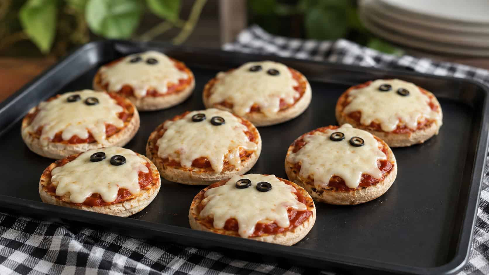 a tray of mini English muffin pizzas each topped with a melty mozzarella ghost and olive eyes in a wide horizontal shot, on a black baking tray over a checkered cloth, natural light, shallow depth of field, landscape orientation