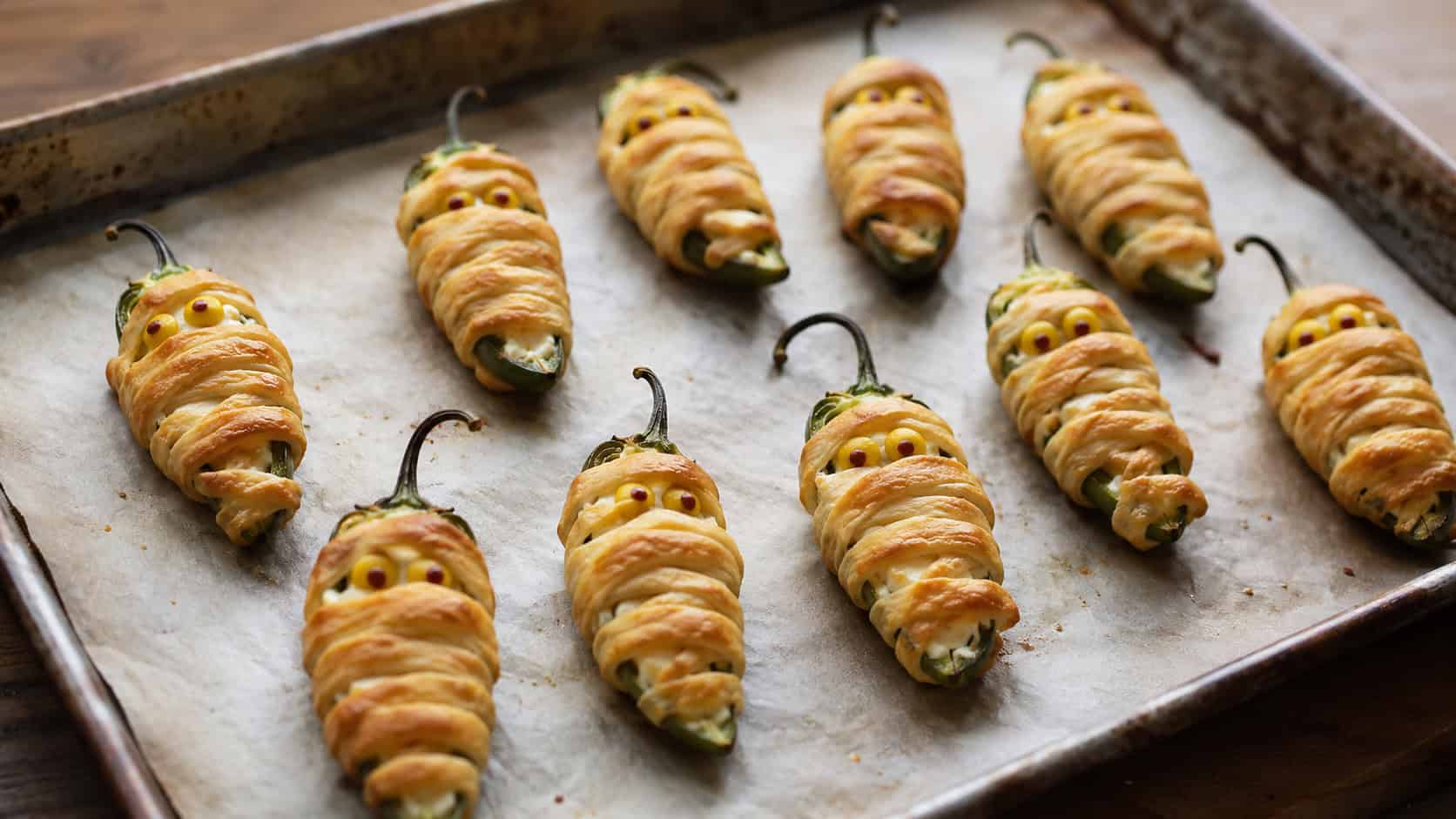 a tray of jalapeno poppers wrapped in golden dough strips like mummies with mustard-dot eyes in a wide horizontal shot, on parchment over a rustic metal sheet pan, natural light, shallow depth of field, landscape orientation