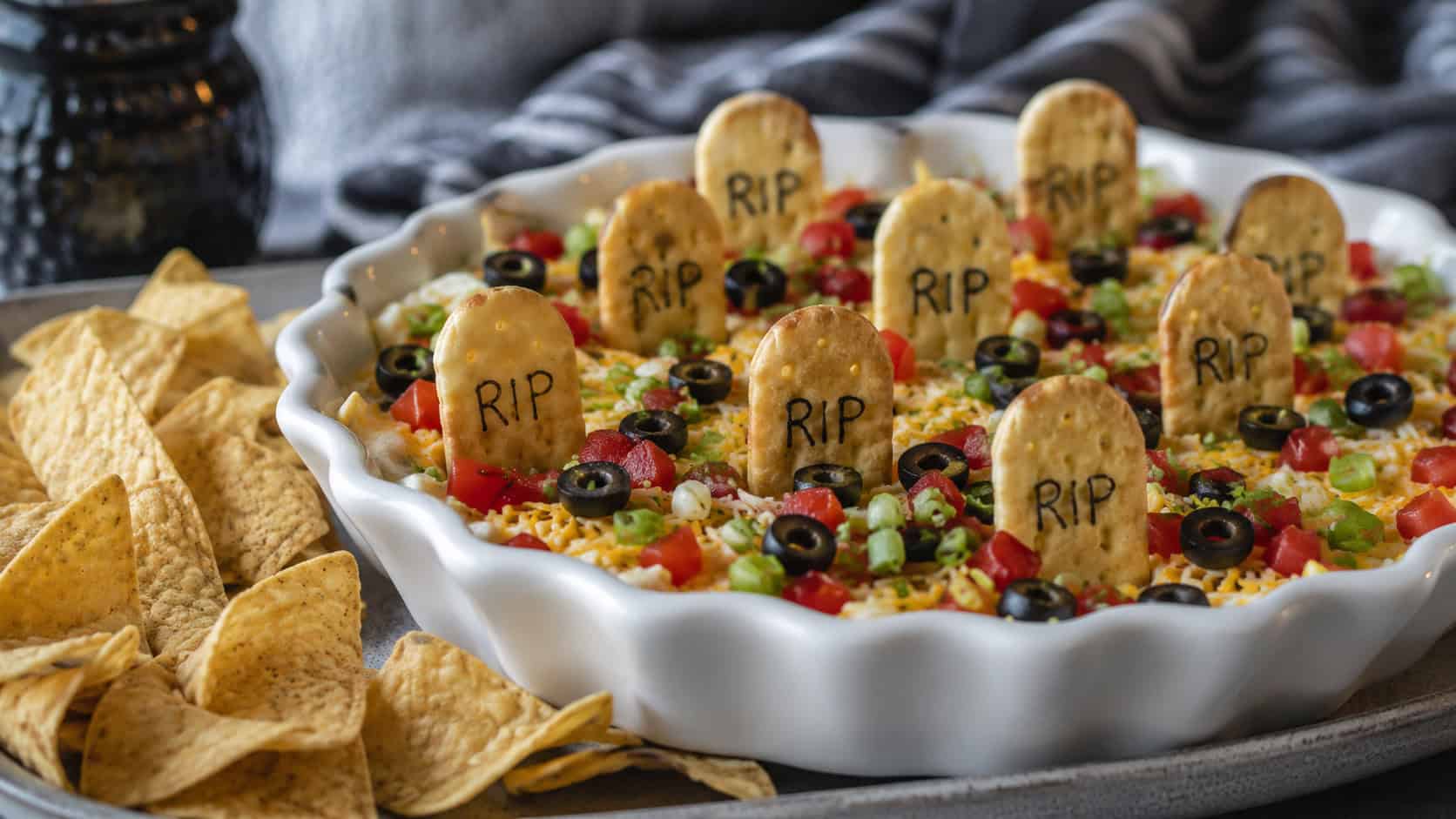 a shallow dish of layered taco dip with cracker tombstones standing up and olive accents in a wide horizontal shot, on a gray platter beside tortilla chips, natural light, shallow depth of field, landscape orientation