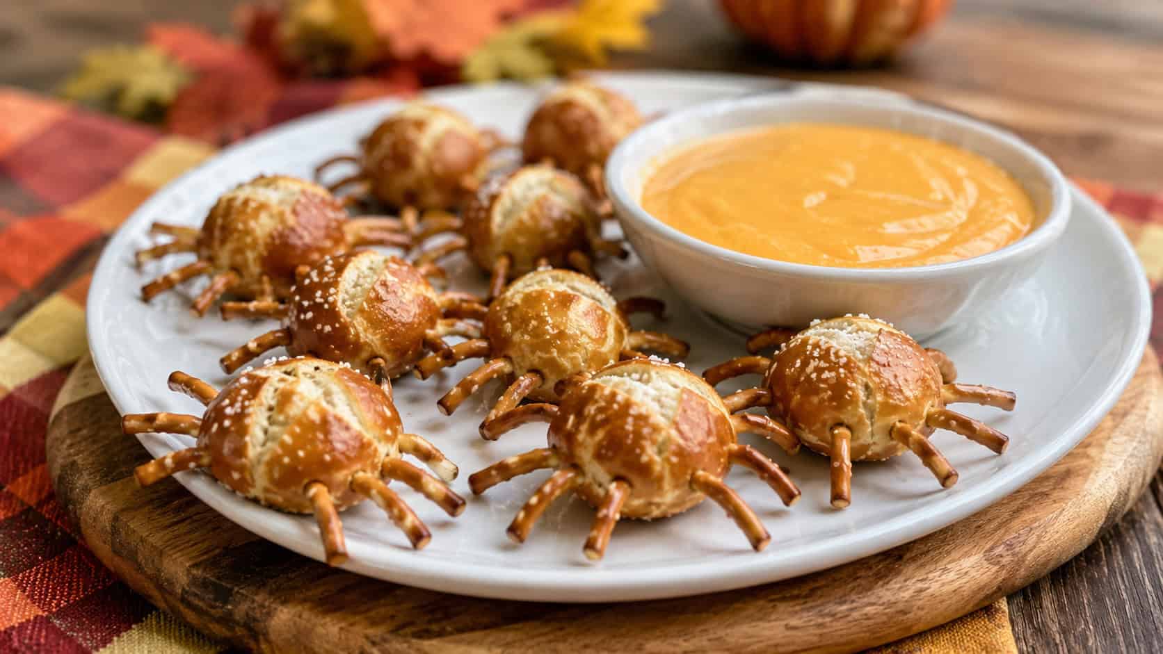 a platter of round soft pretzel bites with thin pretzel-stick legs like spiders beside cheese dip in a wide horizontal shot, on a wooden board over a fall-colored cloth, natural light, shallow depth of field, landscape orientation