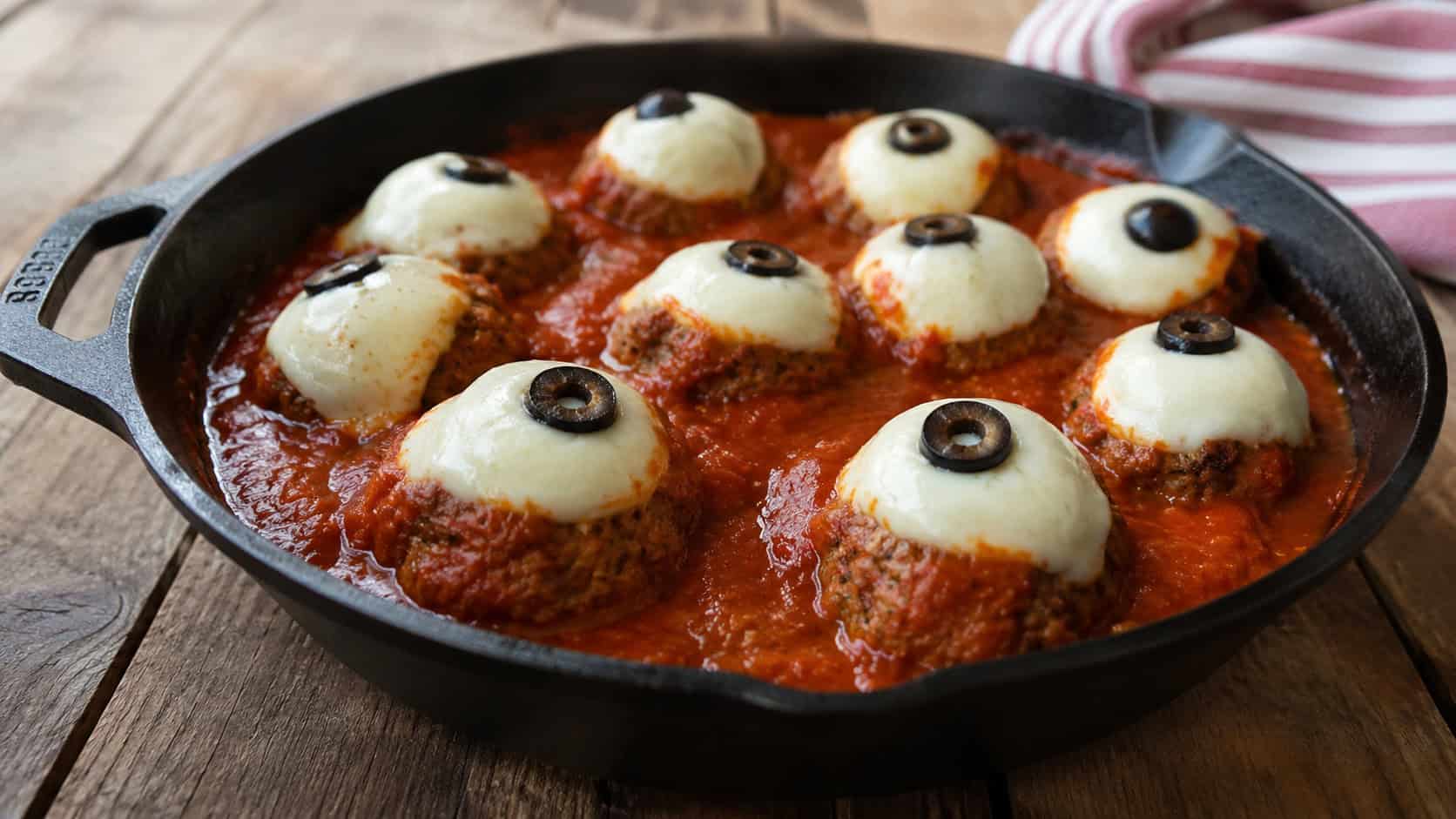 saucy meatballs in red marinara each topped with a mozzarella ball and olive slice like eyeballs in a wide horizontal shot, in a black cast-iron skillet on a wooden table, natural light, shallow depth of field, landscape orientation