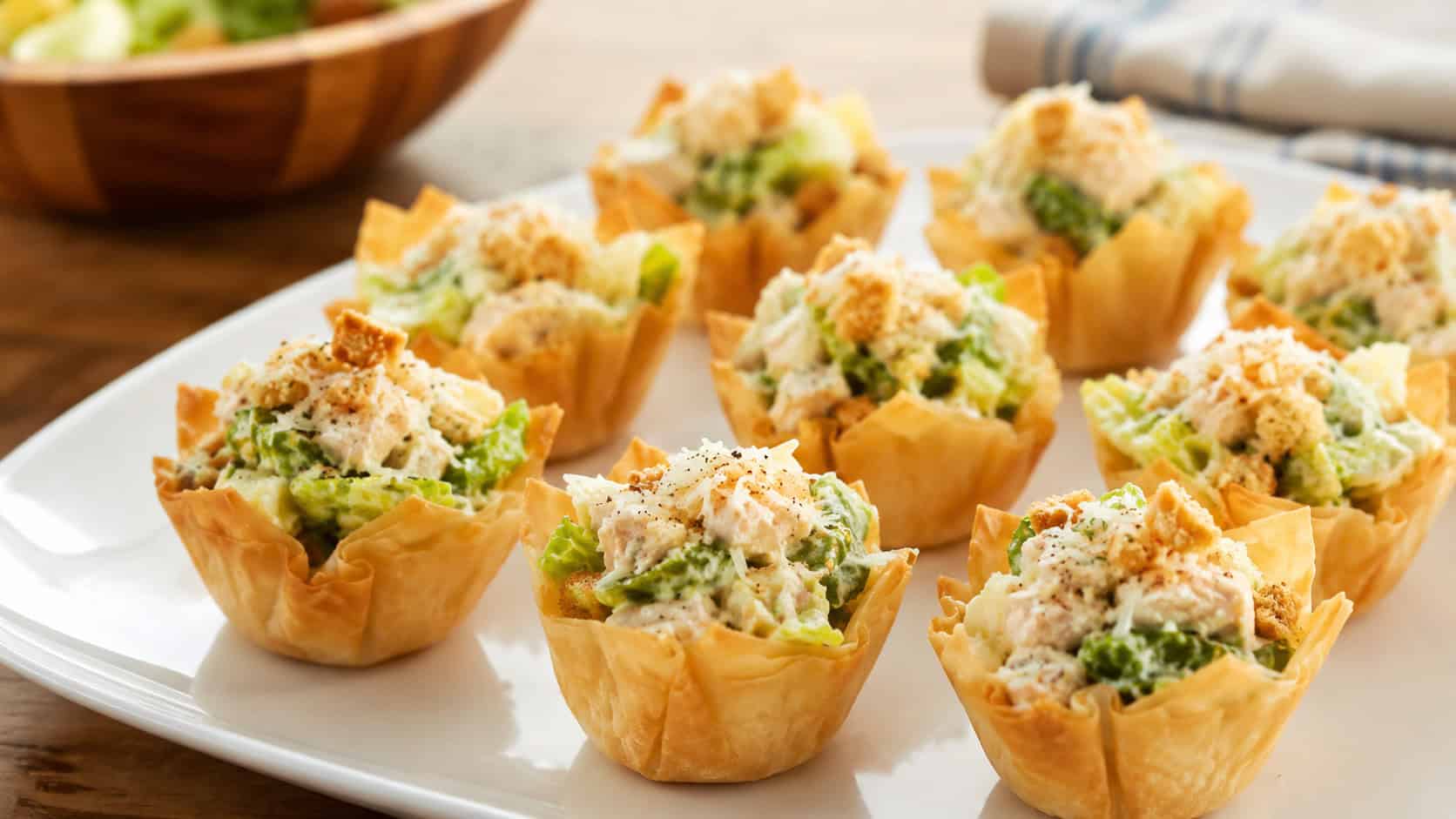 chicken caesar salad phyllo bites on a white plate, golden crisp pastry cups filled with finely shredded green romaine and chopped chicken tossed in creamy dressing, grated parmesan and crushed crouton on top, fresh bright natural light, shallow depth of field, kitchen table