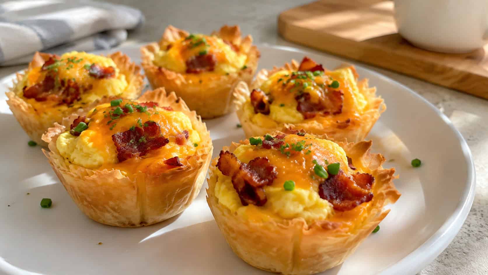 bacon and cheese breakfast phyllo cups on a white plate, golden pastry shells filled with set fluffy egg, melted cheddar and crispy bacon bits, sprinkled with fresh chives, bright morning natural light, shallow depth of field, breakfast kitchen counter