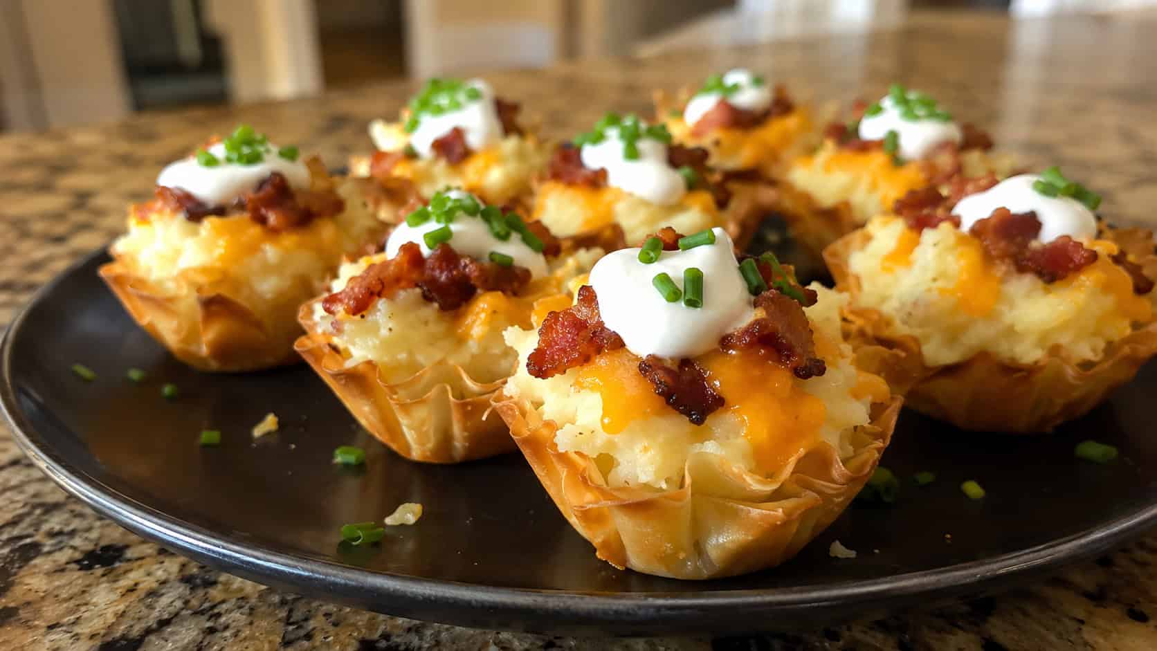 loaded baked potato phyllo bites on a dark plate, golden crisp pastry cups filled with creamy mashed potato topped with melted cheddar, crumbled bacon, a dollop of sour cream and sliced chives, warm natural light, shallow depth of field, cozy kitchen counter