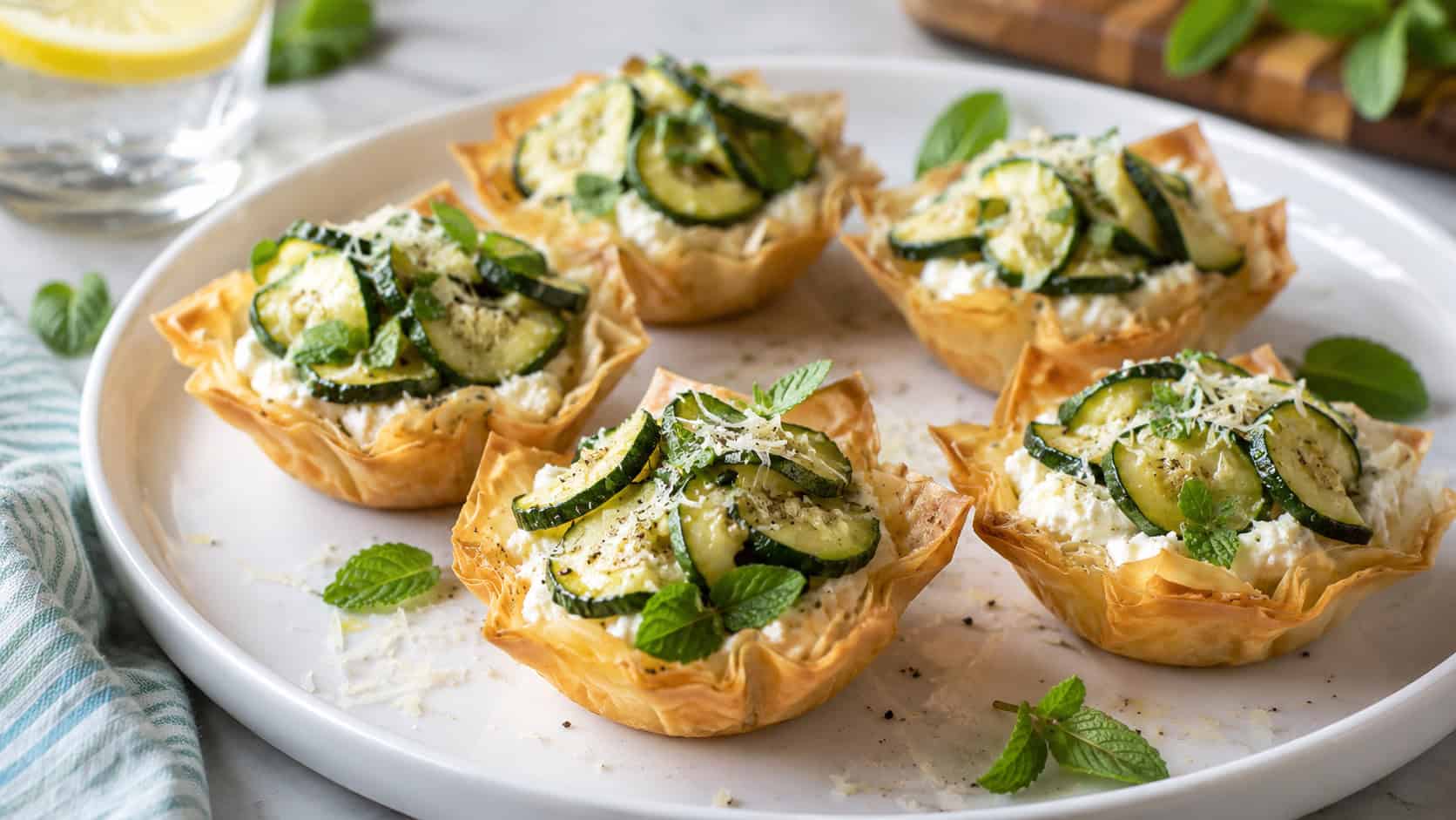zucchini and ricotta phyllo tartlets on a white plate, golden pastry cups filled with creamy white lemon ricotta topped with thin green sautéed zucchini half-moons, scattered fresh mint and parmesan, bright fresh natural light, shallow depth of field, summer kitchen table