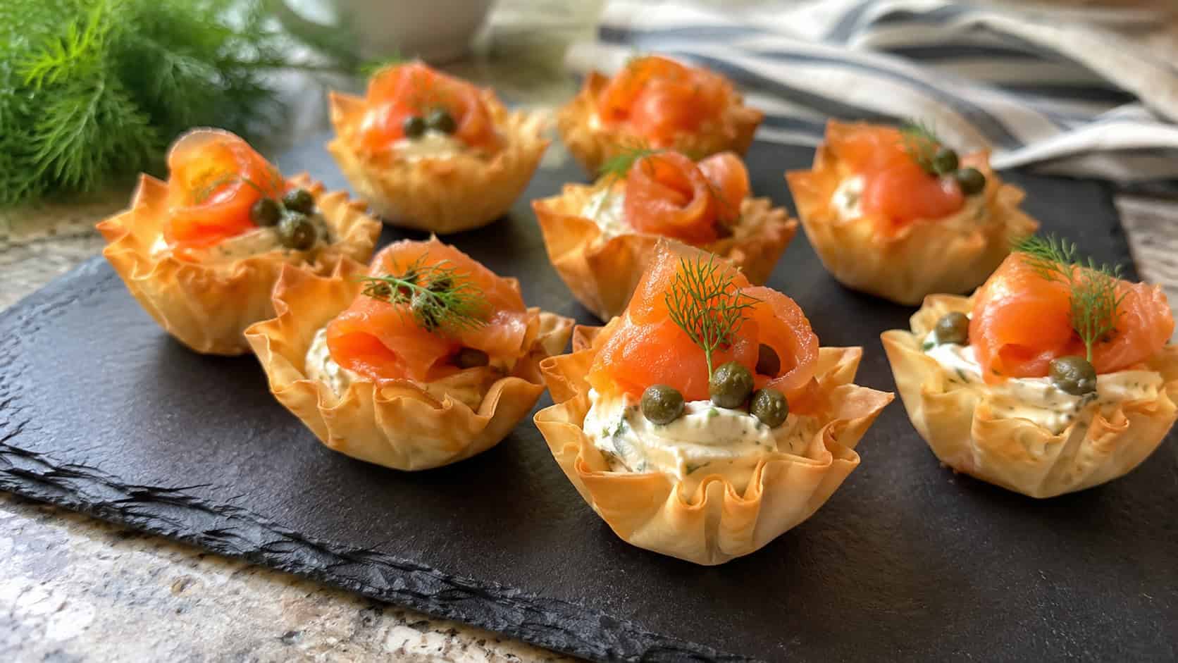 smoked salmon phyllo cups arranged on a slate board, crisp golden pastry filled with swirled herbed cream cheese and a curl of bright orange smoked salmon, tiny capers and fresh dill on top, bright natural light, shallow depth of field, home kitchen setting