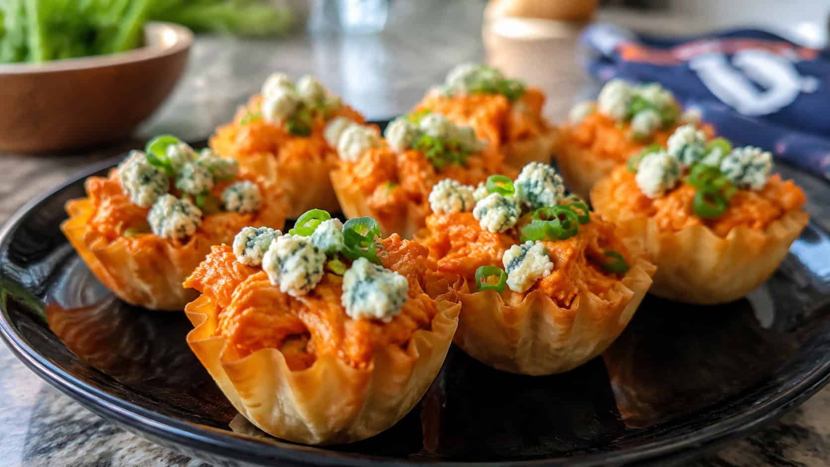 buffalo chicken phyllo cups on a dark plate, crisp golden pastry filled with saucy orange-red shredded buffalo chicken topped with white blue cheese crumbles and sliced green onion, natural light, shallow depth of field, casual kitchen counter, game-day snack vibe
