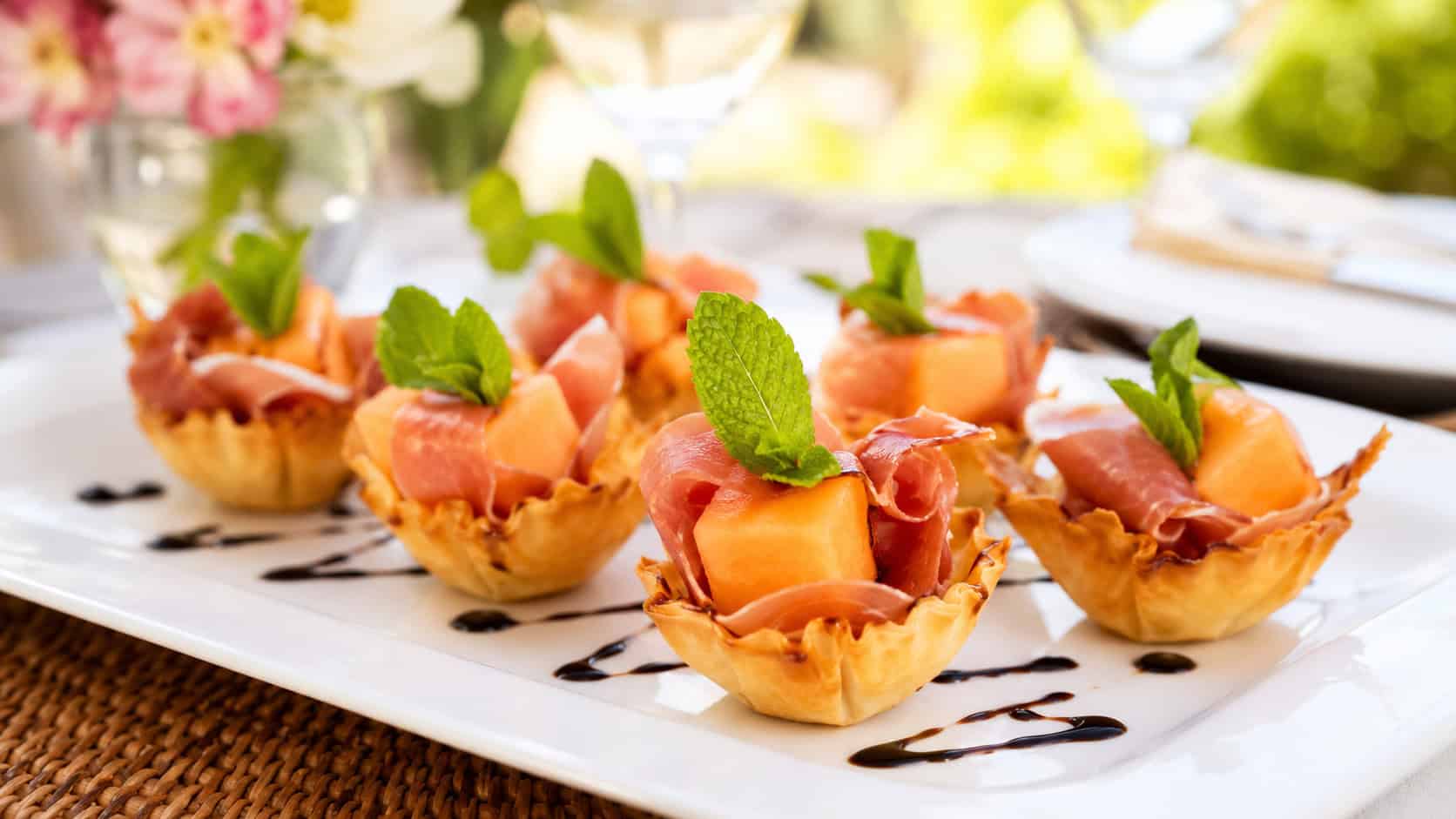 prosciutto and melon phyllo cups on a white plate, golden pastry shells holding orange cantaloupe cubes wrapped with a ruffle of pink prosciutto, fresh green mint leaf on each, a thin balsamic drizzle, bright airy natural light, shallow depth of field, summer table setting