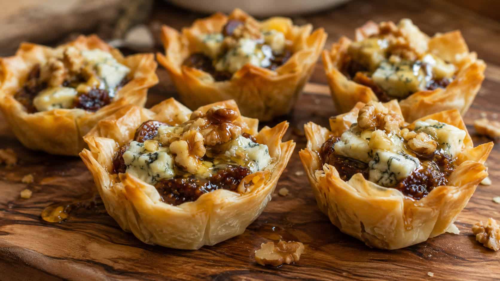 fig and gorgonzola phyllo tartlets on a wooden serving board, golden pastry cups filled with dark fig jam and melting white-blue gorgonzola crumbles, toasted walnut pieces and a thread of honey on top, warm natural light, shallow depth of field, rustic kitchen
