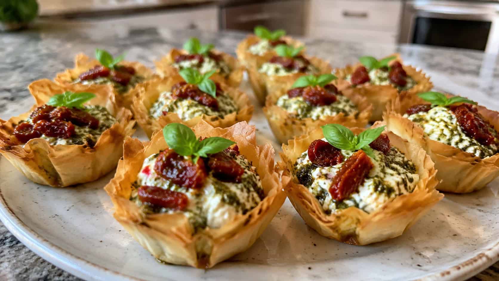 pesto and sun-dried tomato phyllo cups on a ceramic plate, crisp golden shells filled with creamy white ricotta swirled with green basil pesto and deep red chopped sun-dried tomatoes, small basil leaf on top, natural light, shallow depth of field, home kitchen counter
