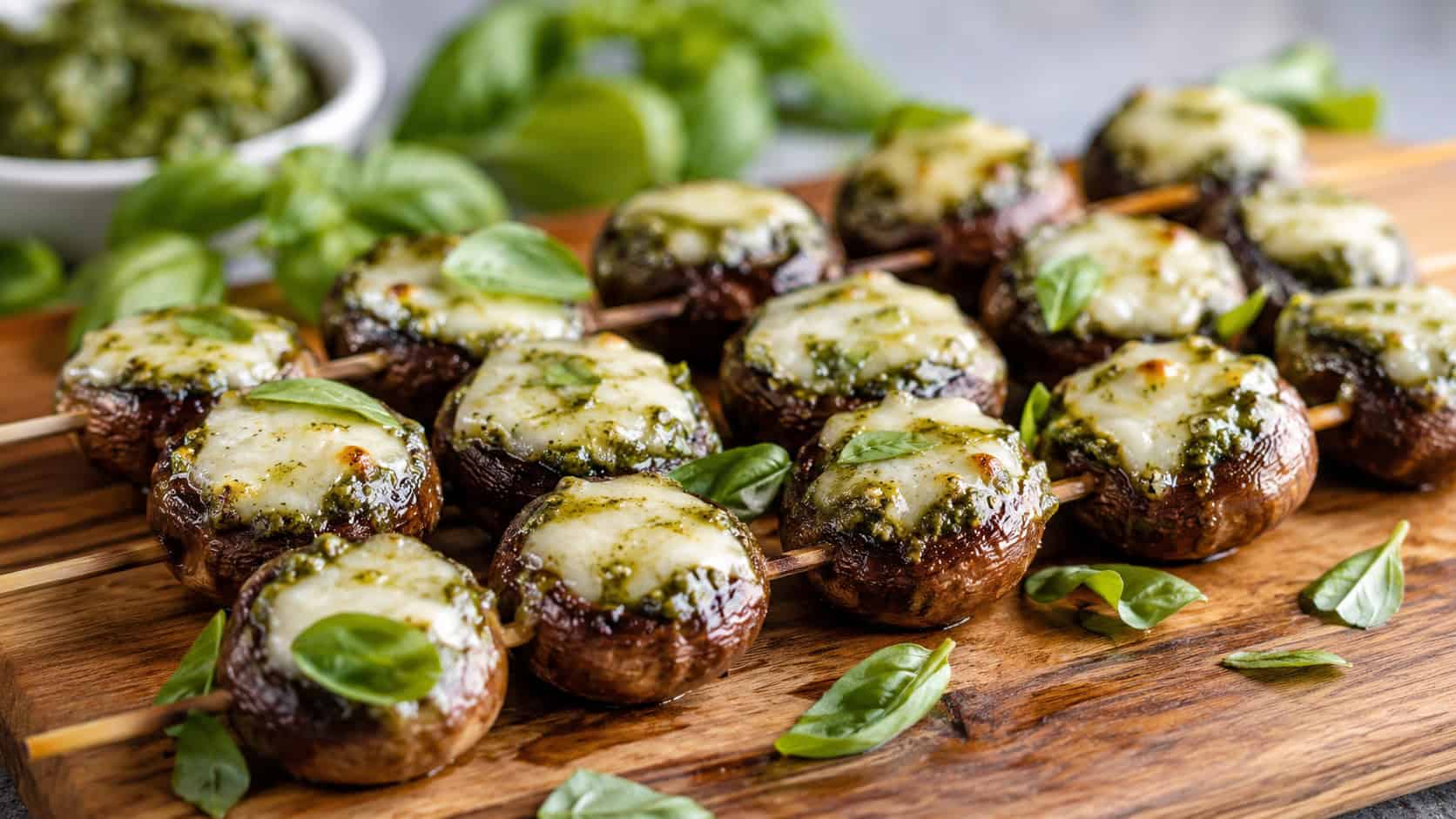 pesto and mozzarella stuffed mushroom skewers on a wooden board in a wide horizontal shot, glistening caps with melty cheese and green pesto, fresh basil scattered around, soft natural light, shallow depth of field, landscape orientation