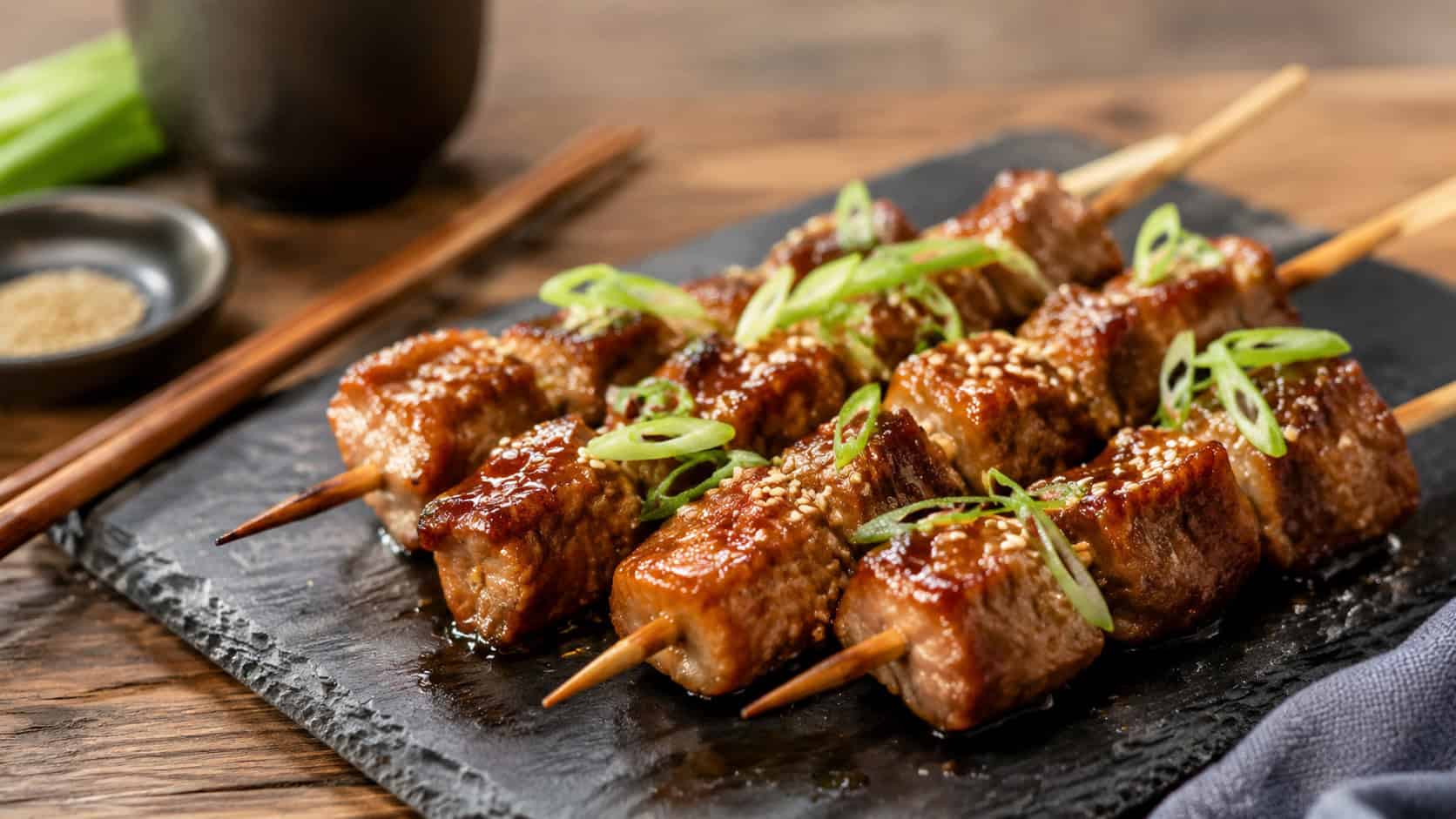 soy ginger glazed pork skewers on a slate plate in a wide horizontal shot, glossy caramelized cubes, sesame seeds and scallion slivers on top, chopsticks resting nearby, warm natural light, shallow depth of field, landscape orientation
