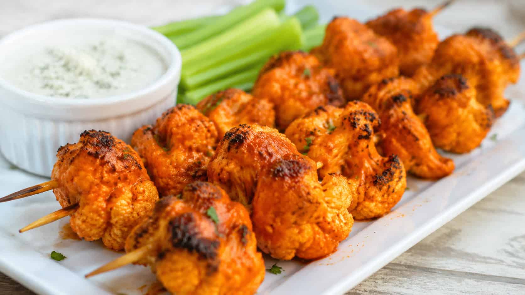 buffalo cauliflower skewers on a white platter in a wide horizontal shot, fiery orange-coated florets with charred edges, celery sticks and a ramekin of blue cheese dressing alongside, bright natural light, shallow depth of field, landscape orientation