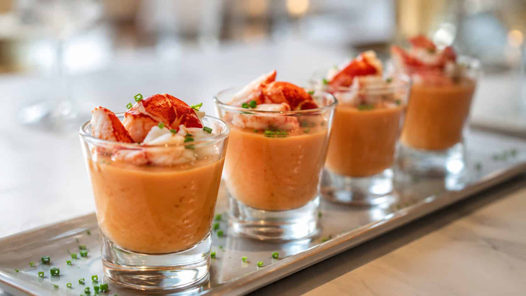 small glass shooters of creamy orange lobster bisque garnished with lobster meat in a wide horizontal shot, lined up on a sleek tray with a sprinkle of chives, soft elegant light, shallow depth of field, landscape orientation