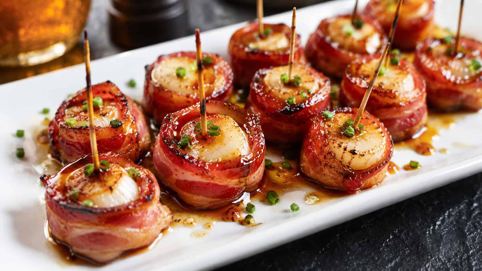 bacon-wrapped scallops with a glossy maple glaze on a platter in a wide horizontal shot, garnished with chives on a dark board with toothpicks, warm appetizing light, shallow depth of field, landscape orientation