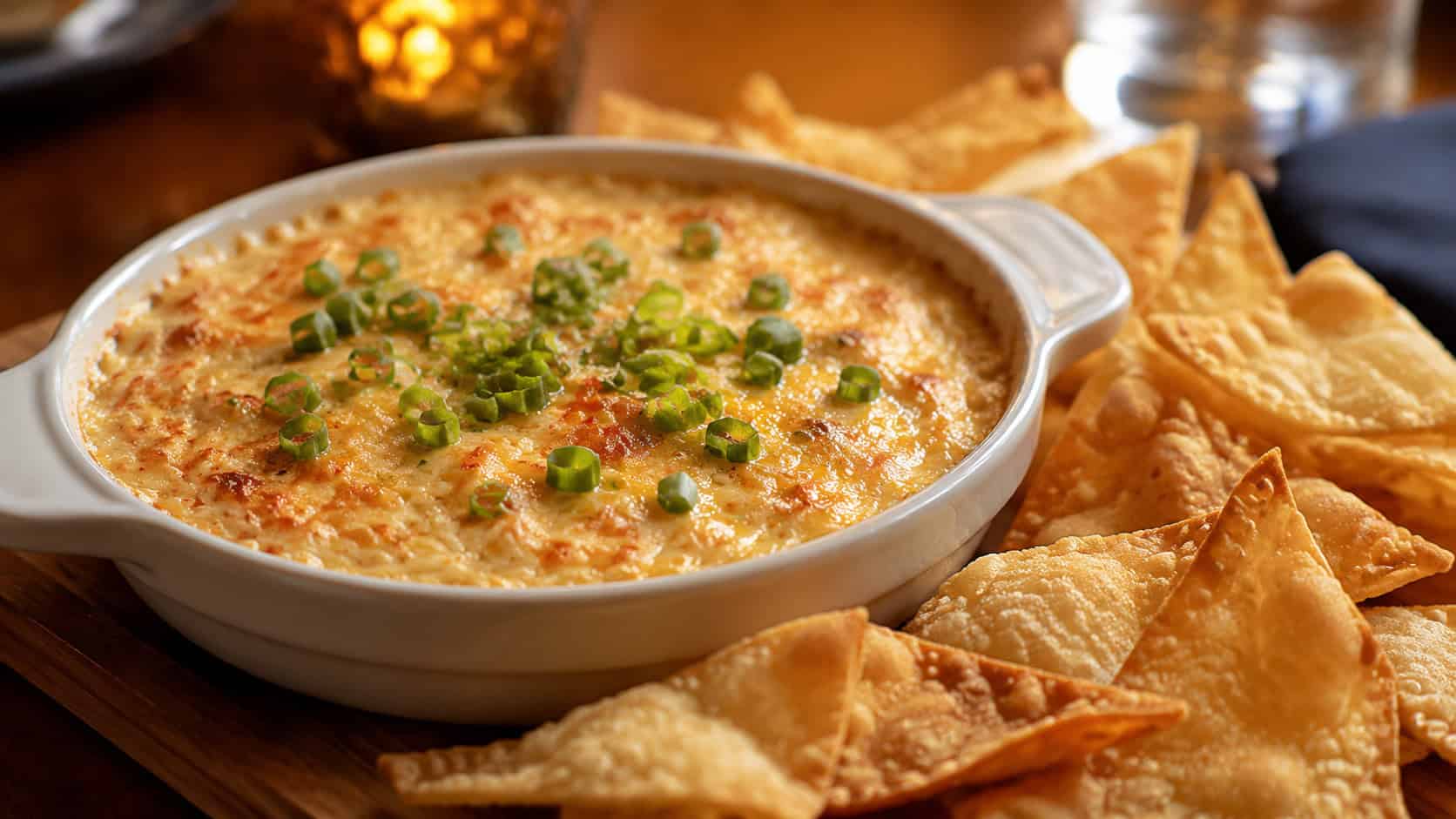a bubbling golden crab dip in a ceramic dish surrounded by crispy wonton chips in a wide horizontal shot, on a wooden serving board with green onion garnish, warm cozy light, shallow depth of field, landscape orientation