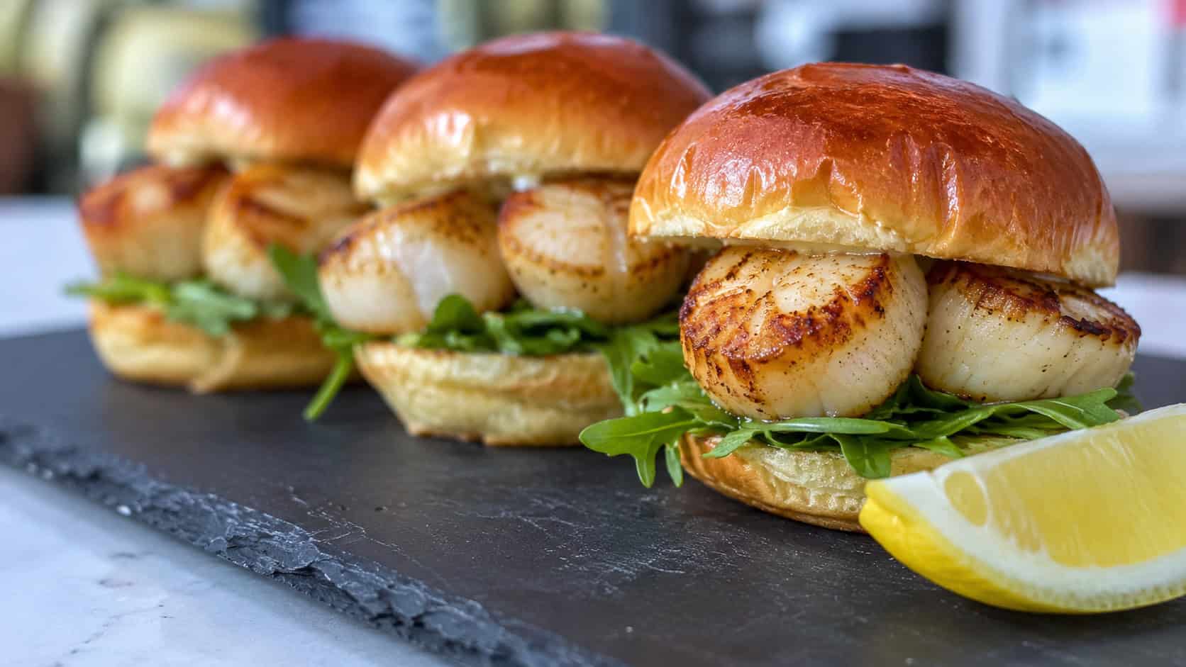 golden seared scallop sliders on toasted brioche buns with arugula peeking out in a wide horizontal shot, on a slate board with a lemon wedge beside them, bright natural light, shallow depth of field, landscape orientation