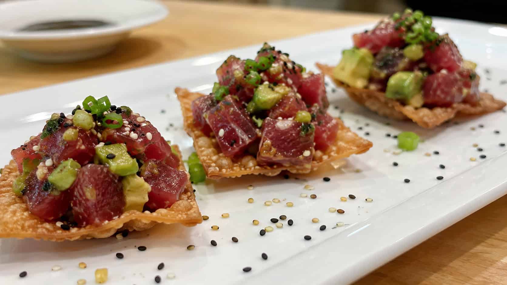 ruby ahi tuna tartare with avocado spooned onto crisp wonton chips in a wide horizontal shot, on a white platter with sesame seeds scattered around, clean bright light, shallow depth of field, landscape orientation