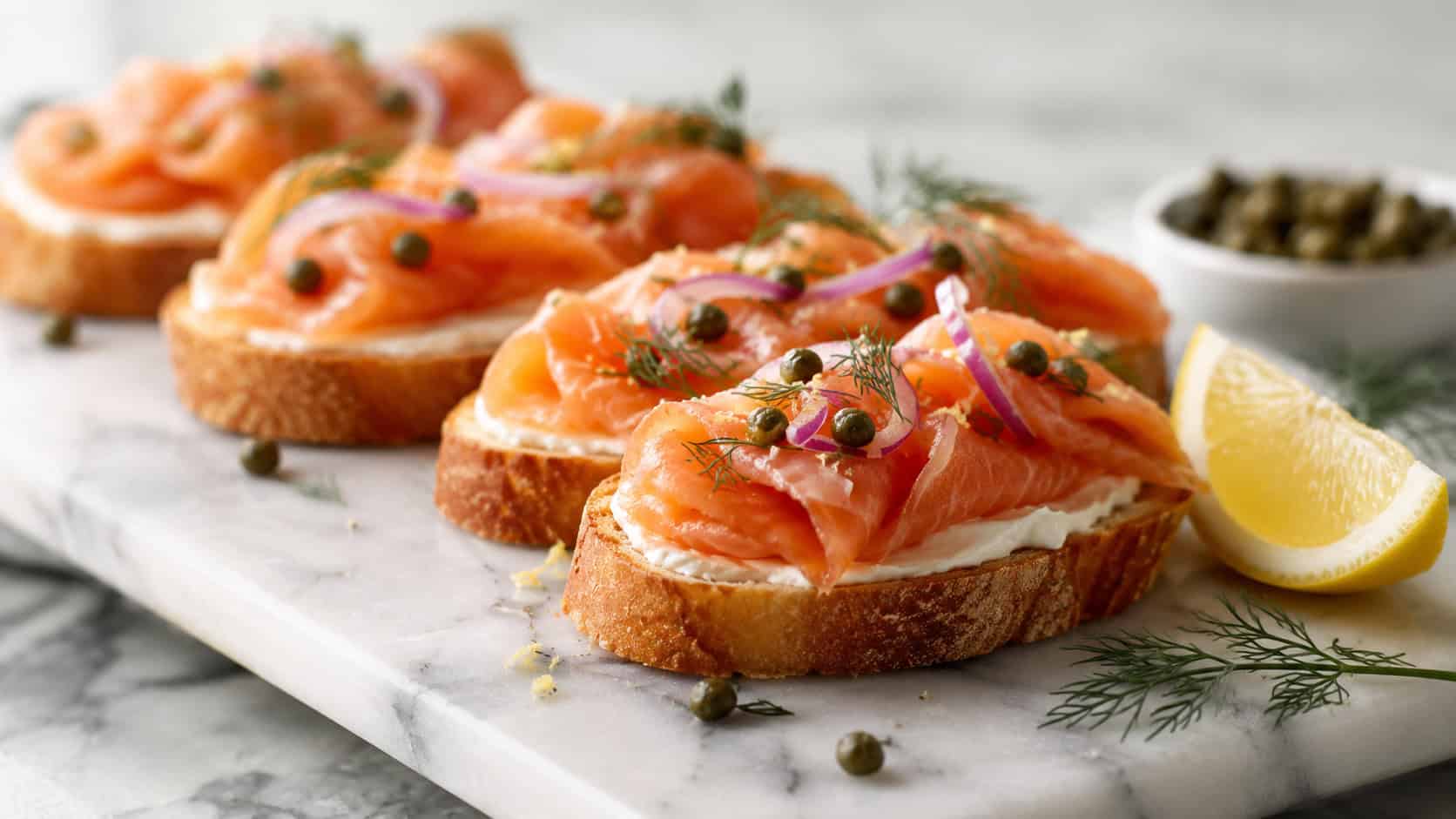 smoked salmon crostini topped with capers and red onion in a wide horizontal shot, lined on a marble board with a lemon and dill sprigs nearby, fresh bright light, shallow depth of field, landscape orientation