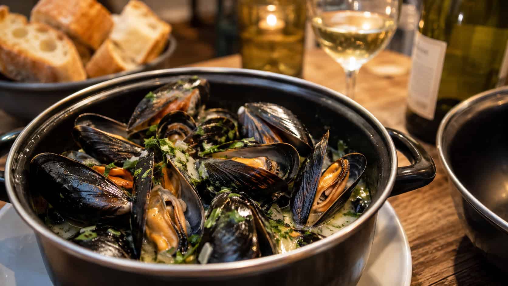 an open pot of steamed mussels in a glossy white wine broth with parsley in a wide horizontal shot, on a rustic table with crusty bread and wine alongside, warm bistro light, shallow depth of field, landscape orientation