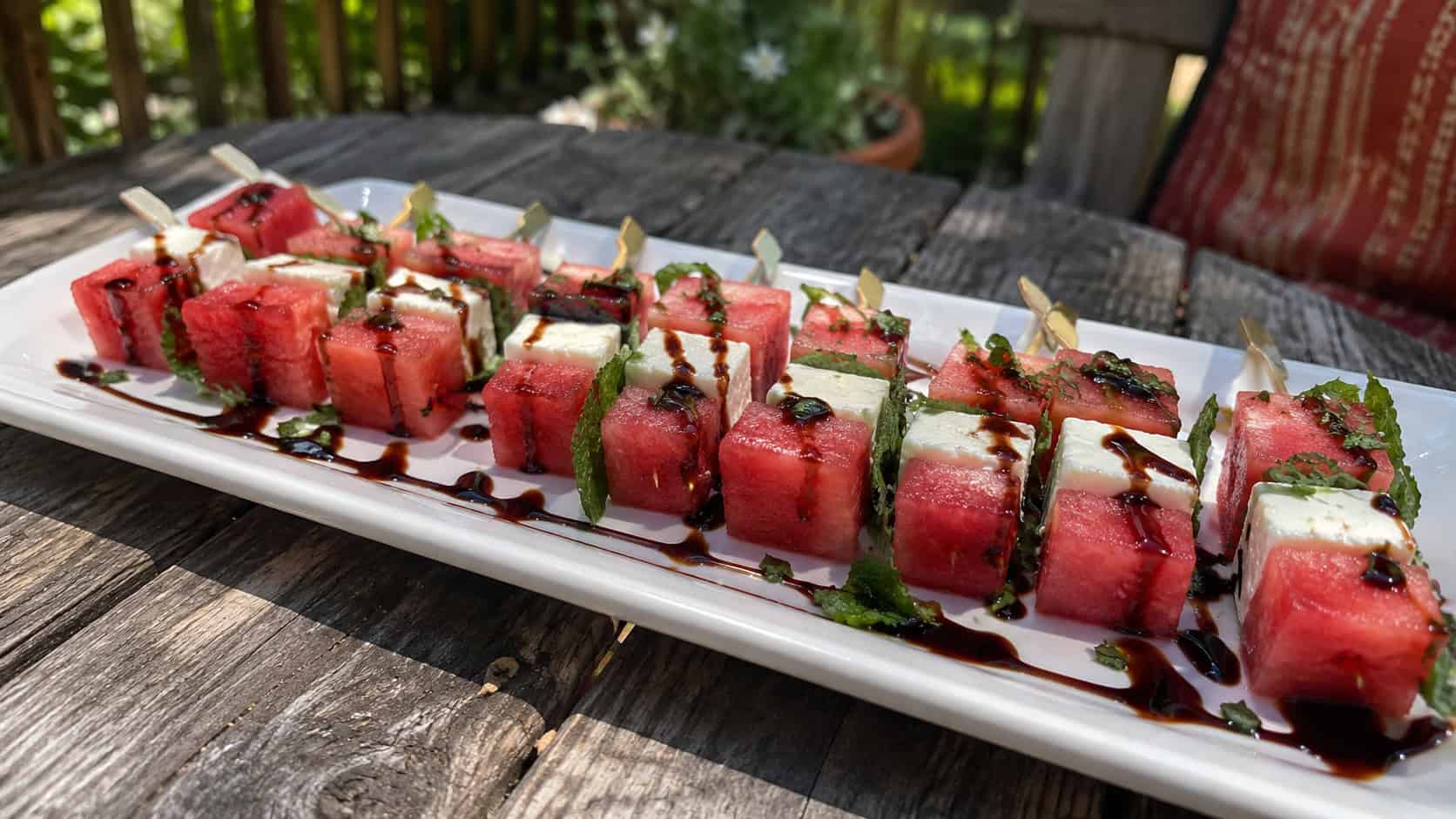 a long white platter of watermelon and feta mint skewers in a wide horizontal shot, drizzled with balsamic glaze on a weathered wooden patio table, natural sunlight, shallow depth of field, landscape orientation