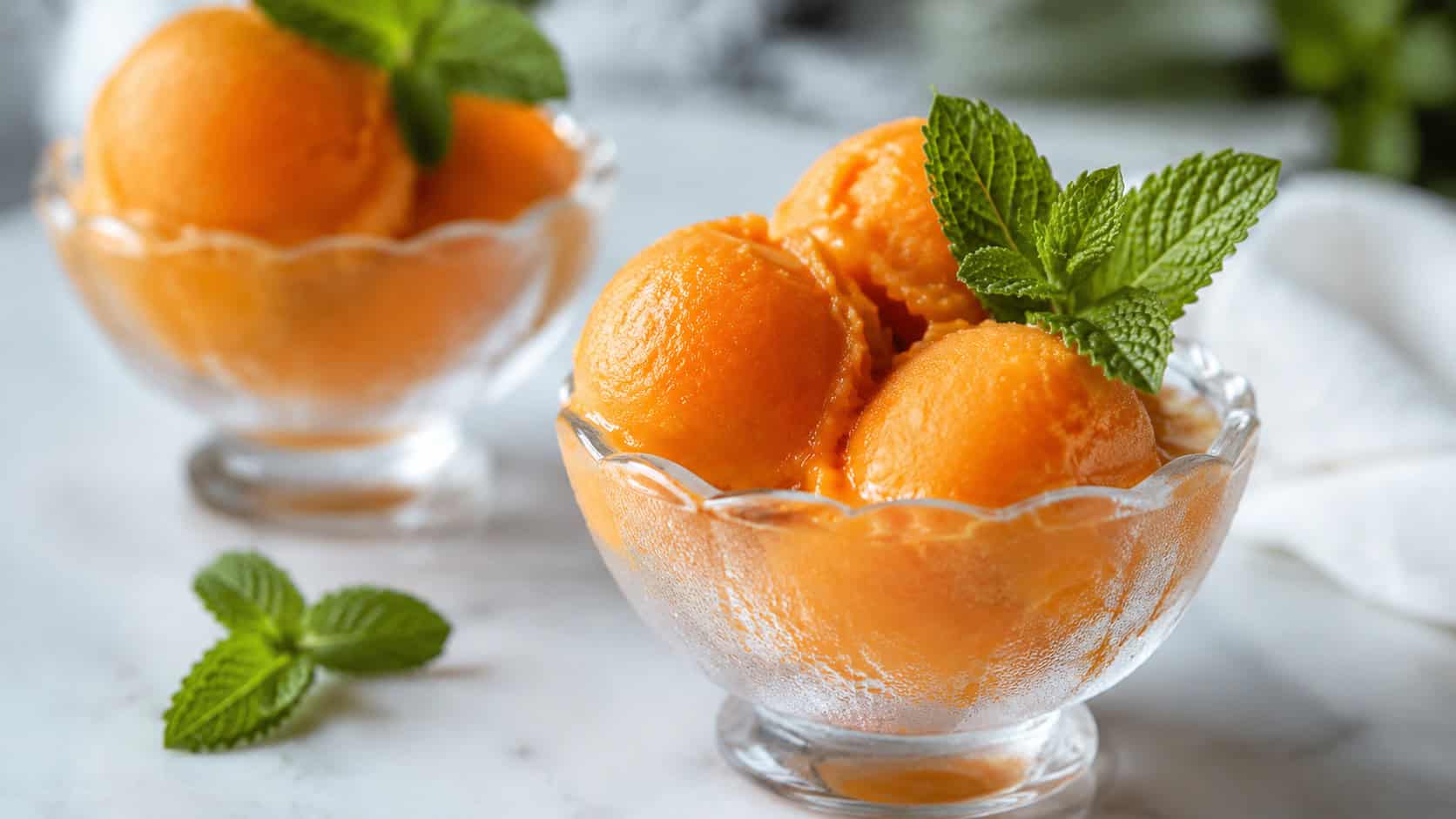 scoops of bright orange sorbet garnished with mint in a wide horizontal shot, served in chilled glass bowls on a white table, cool natural light, shallow depth of field, landscape orientation