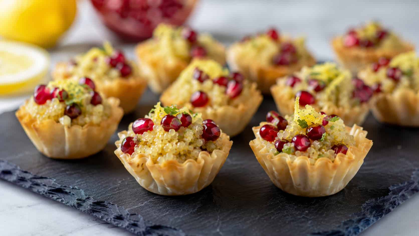 mini phyllo cups filled with lemony quinoa and ruby pomegranate seeds in a wide horizontal shot, arranged on a dark slate platter, bright natural light, shallow depth of field, landscape orientation