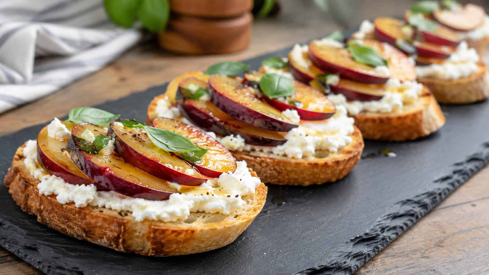 bruschetta topped with ricotta, sliced plums, and basil in a wide horizontal shot, lined up on a long slate board, bright natural light, shallow depth of field, landscape orientation