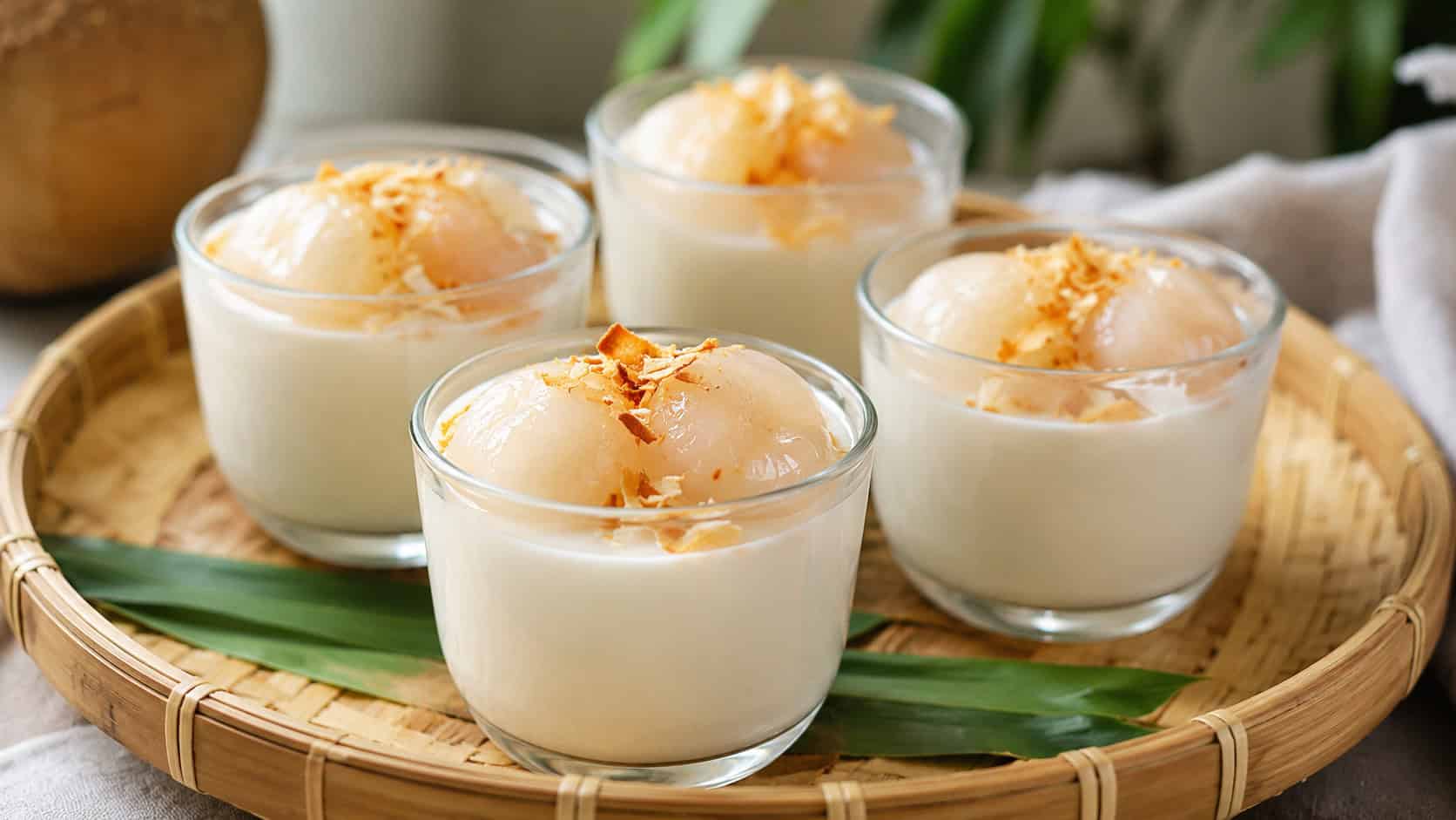 small glass cups of creamy coconut pudding topped with lychee and toasted coconut in a wide horizontal shot, on a bamboo tray, soft natural light, shallow depth of field, landscape orientation