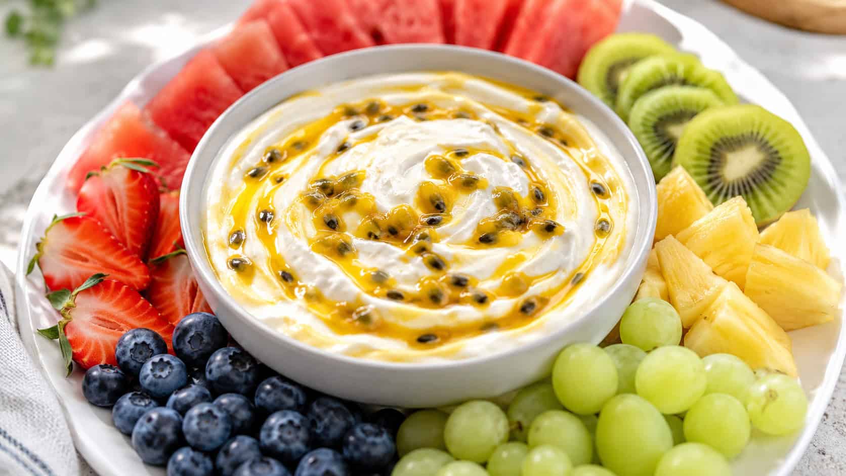 a bowl of creamy yogurt dip swirled with golden passion fruit in a wide horizontal shot, surrounded by colorful fresh fruit for dipping on a white platter, bright natural light, shallow depth of field, landscape orientation