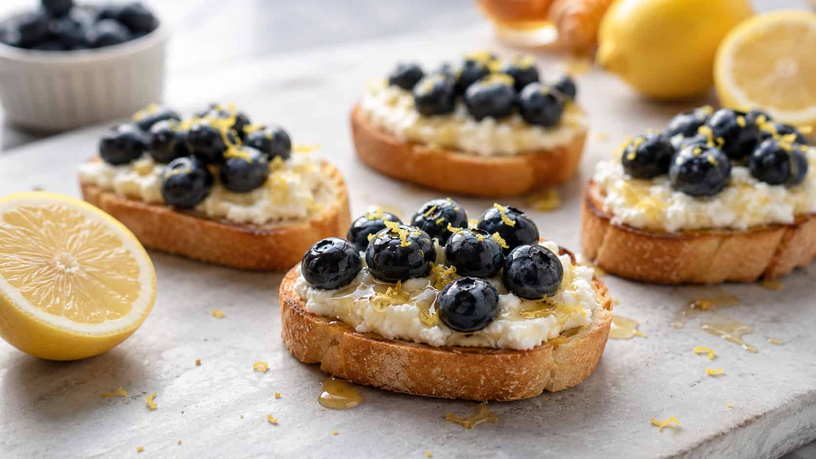 lemon ricotta crostini topped with fresh blueberries and honey in a wide horizontal shot, laid out on a light gray board with lemon halves, bright natural light, shallow depth of field, landscape orientation