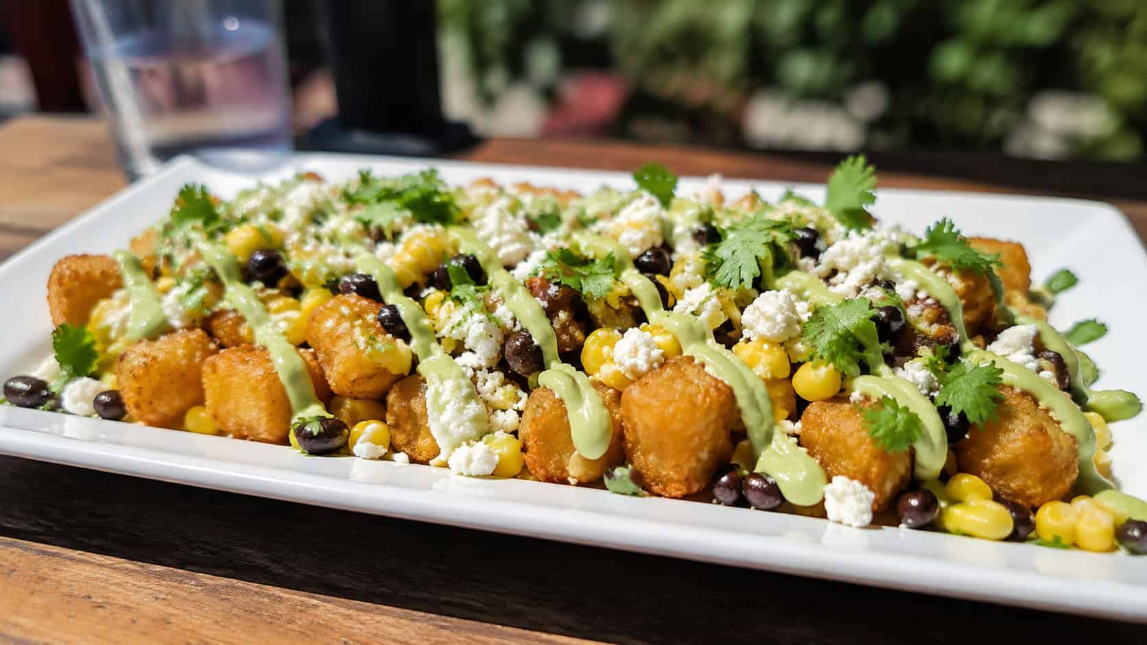 southwest tater tots topped with corn, black beans, and crumbled cotija in a wide horizontal shot, a lime crema drizzle and cilantro across the top, bright natural light, shallow depth of field, landscape orientation