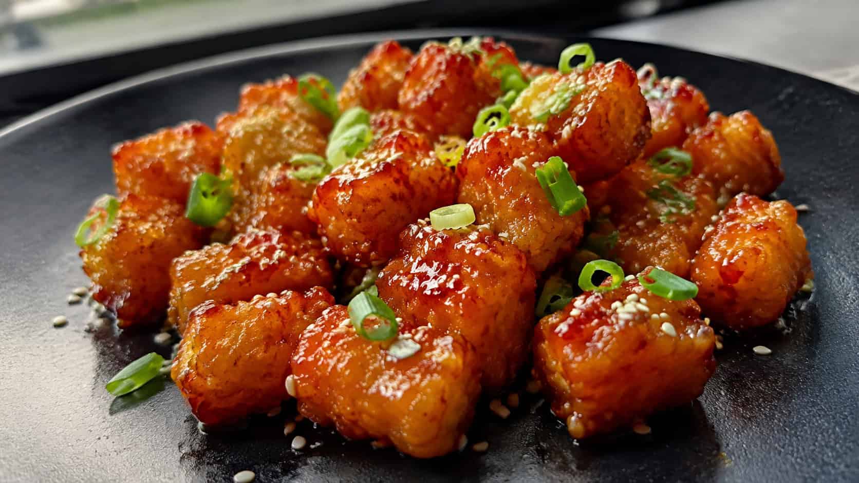 glossy sriracha-glazed tater tots sprinkled with sesame seeds and green onions in a wide horizontal shot on a dark plate, the red glaze catching the light, natural light, shallow depth of field, landscape orientation