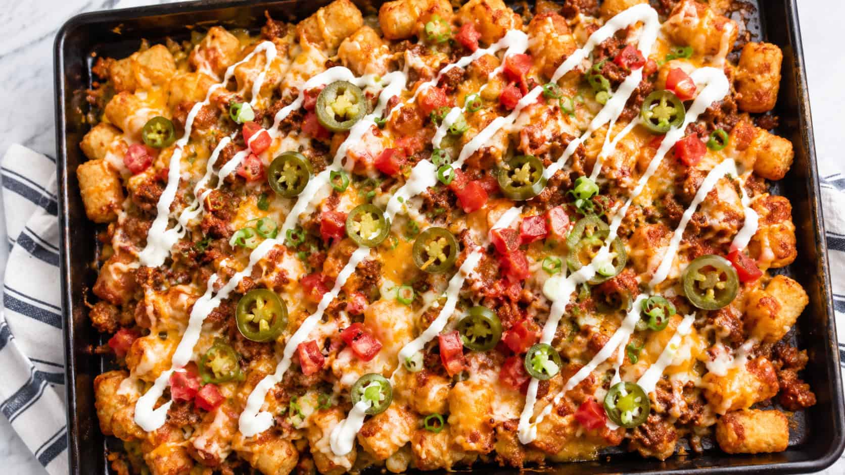 a loaded sheet pan of tater tot nachos covered in melted cheese, seasoned beef, diced tomato, green onion, and jalapenos in a wide horizontal overhead shot, a drizzle of sour cream on top, bright natural light, shallow depth of field, landscape orientation