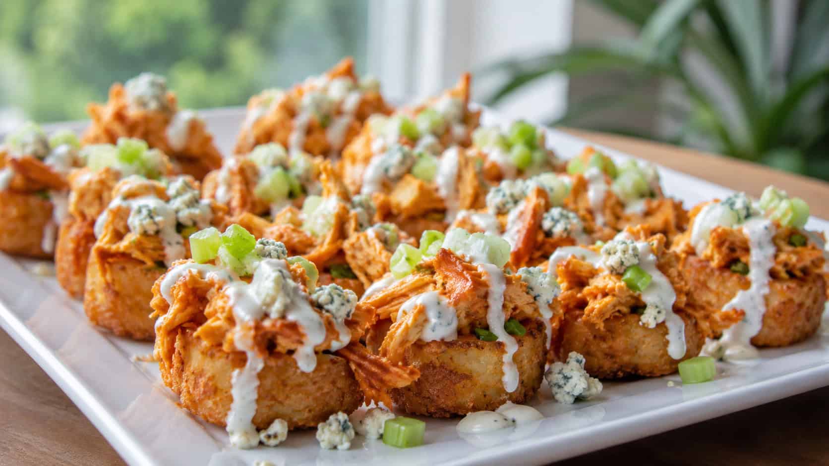 buffalo chicken tater tot bites topped with orange buffalo chicken, blue cheese crumbles, and diced celery in a wide horizontal shot on a white platter, a ranch drizzle across the top, natural light, shallow depth of field, landscape orientation