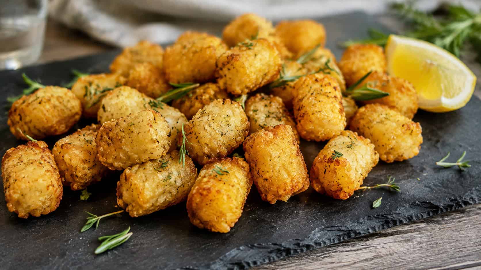 golden garlic herb tater tots scattered with fresh rosemary and thyme in a wide horizontal shot on a slate serving board, a wedge of lemon to the side, bright natural light, shallow depth of field, landscape orientation