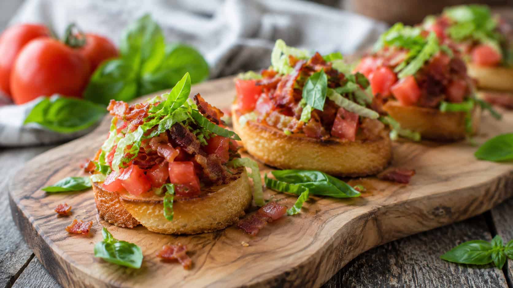 BLT bruschetta arranged on a wide wooden serving board shot horizontally, crisp toast rounds heaped with diced red tomato, crumbled bacon and shredded lettuce, fresh basil and a rustic surface, bright natural light, shallow depth of field, landscape orientation