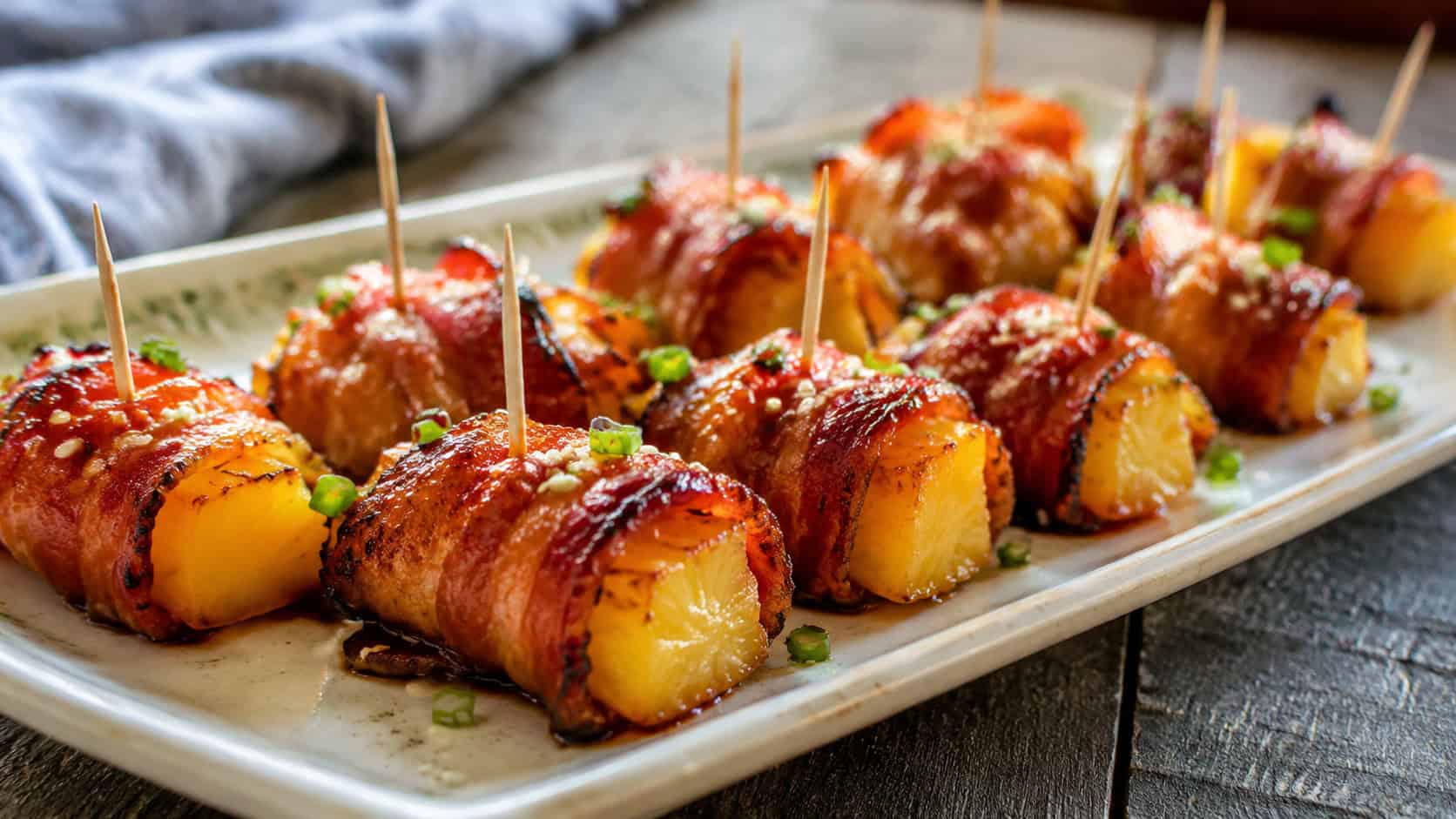bacon-wrapped pineapple bites on a wide ceramic platter shot horizontally, glossy glazed bacon around golden pineapple with toothpicks, a sprinkle of sesame and green onion, natural light, shallow depth of field, landscape orientation