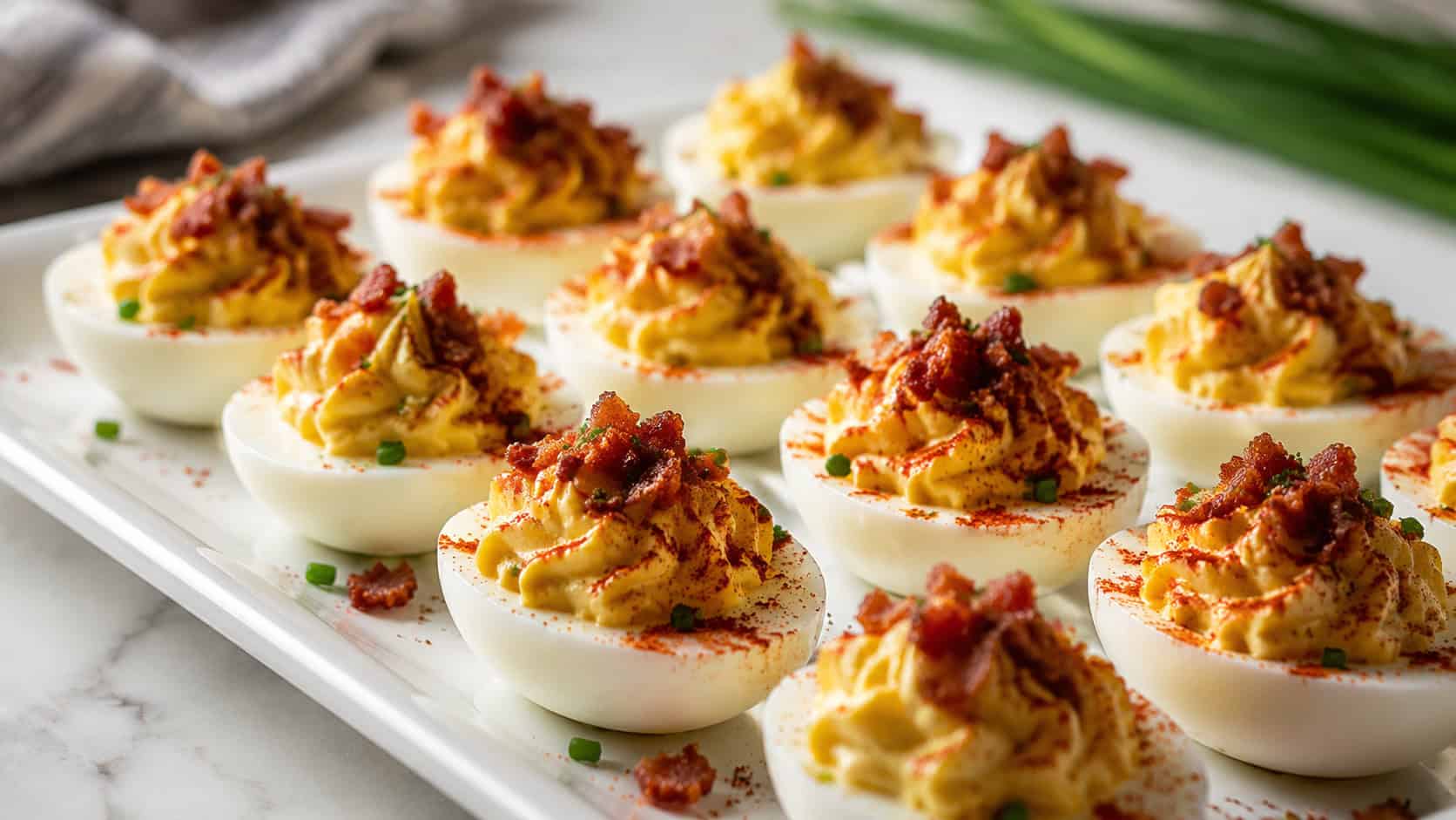 smoky bacon deviled eggs on a wide white deviled egg platter shot horizontally, creamy piped yolk filling dusted with paprika and topped with crumbled bacon, fresh chives and a light surface, bright natural light, shallow depth of field, landscape orientation