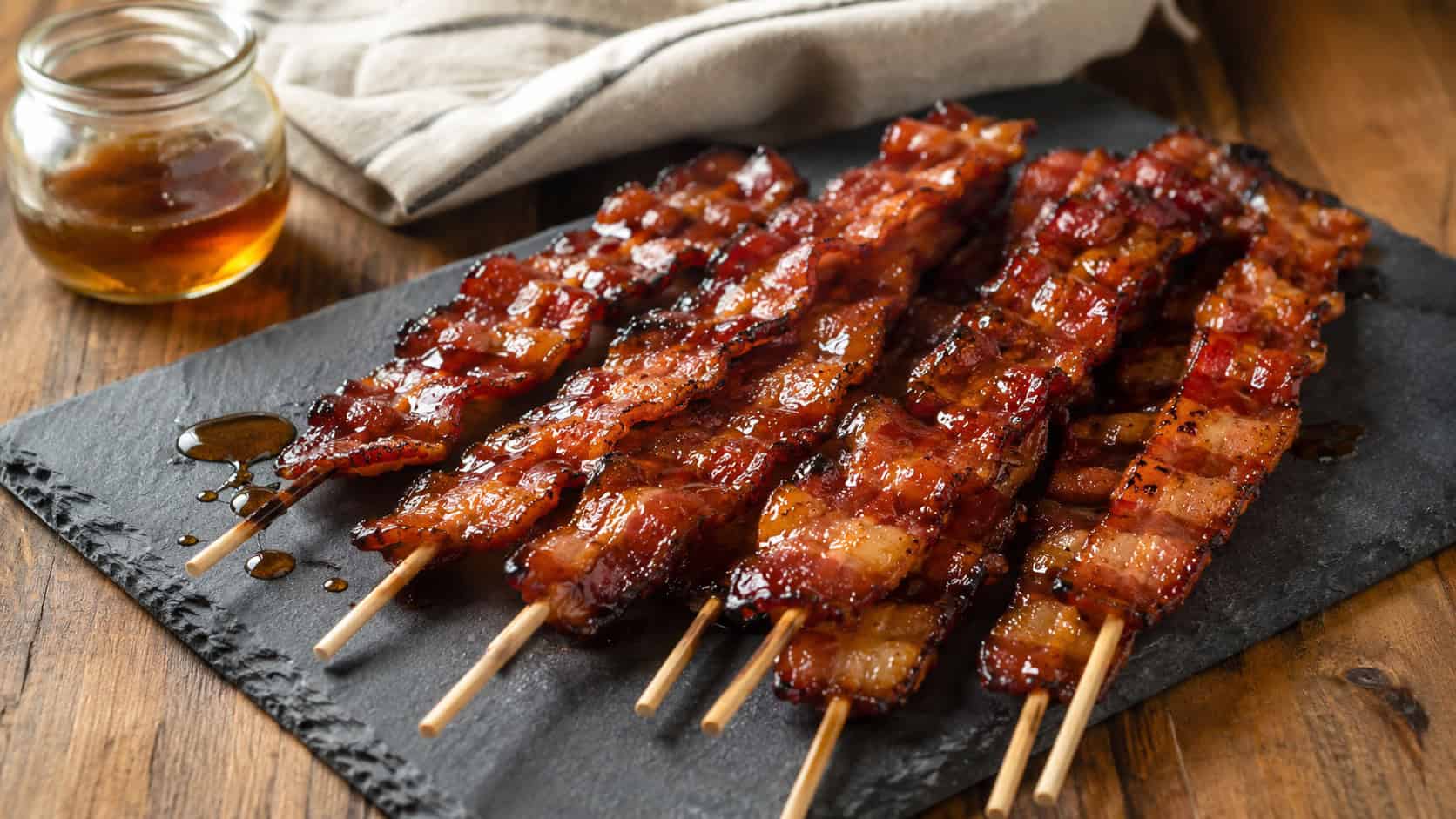 maple-glazed bacon skewers fanned across a slate board in a wide horizontal shot, glossy caramelized bacon on wooden sticks, a drizzle of maple syrup and a linen napkin nearby, warm natural light, shallow depth of field, landscape orientation