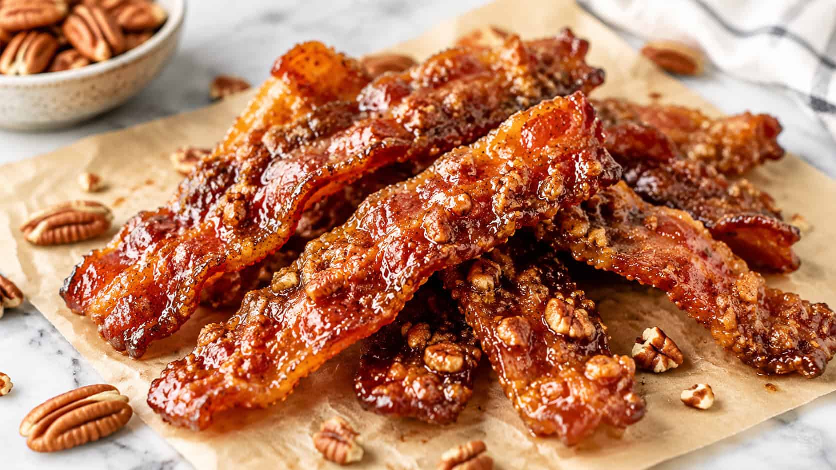 candied pecan bacon strips piled on parchment in a wide horizontal shot, glossy amber bacon studded with toasted pecans, scattered nuts and a marble surface, bright natural light, shallow depth of field, landscape orientation