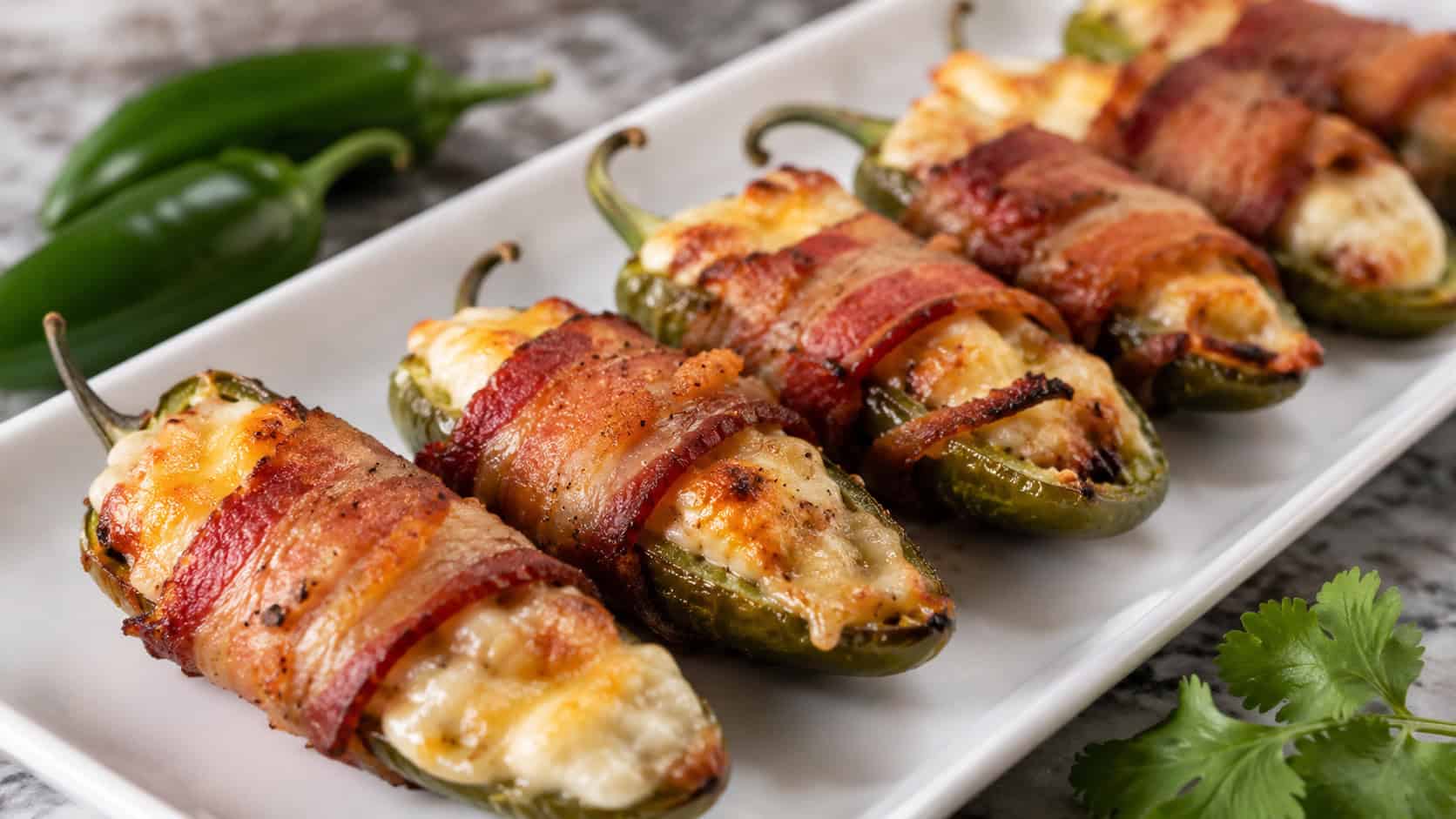 bacon-wrapped jalapeño poppers lined up on a wide white serving board in a horizontal shot, crisp bacon and oozing melted cheese, fresh jalapeños and a cilantro sprig nearby, natural light, shallow depth of field, landscape orientation