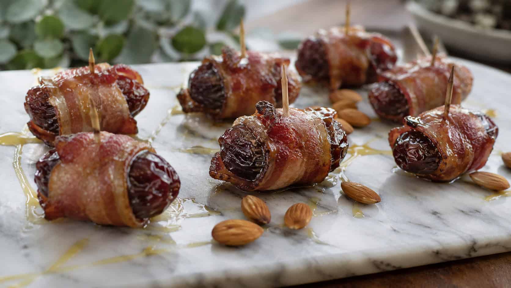 bacon-wrapped dates arranged on a wide marble board in a horizontal shot, glossy caramelized bacon around plump dark dates with toothpicks, a drizzle of honey and scattered almonds, natural light, shallow depth of field, landscape orientation