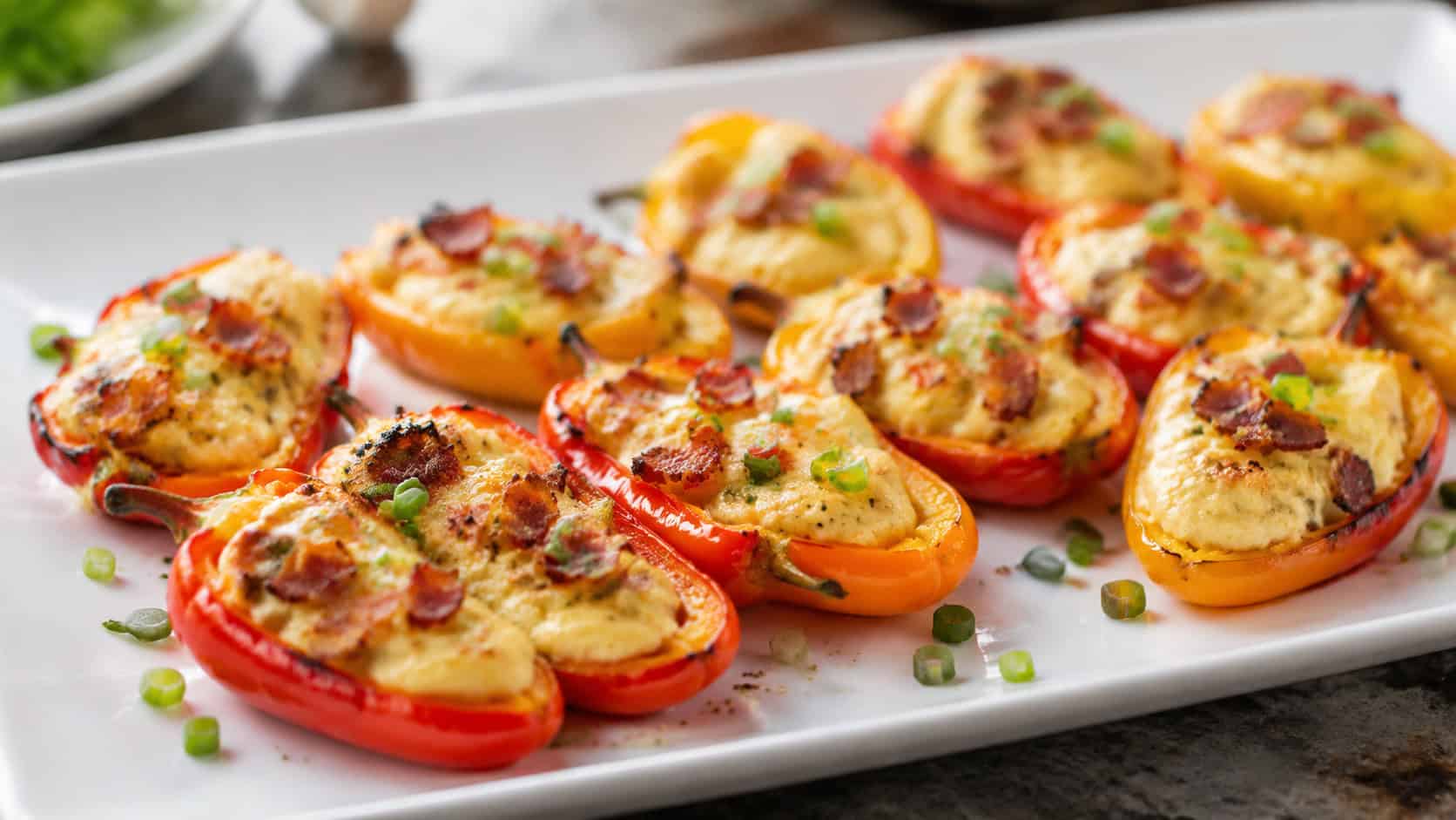 bacon and cheddar stuffed mini peppers on a wide white platter shot horizontally, colorful red yellow and orange peppers with bubbly cheese filling, crumbled bacon and green onion garnish, bright natural light, shallow depth of field, landscape orientation