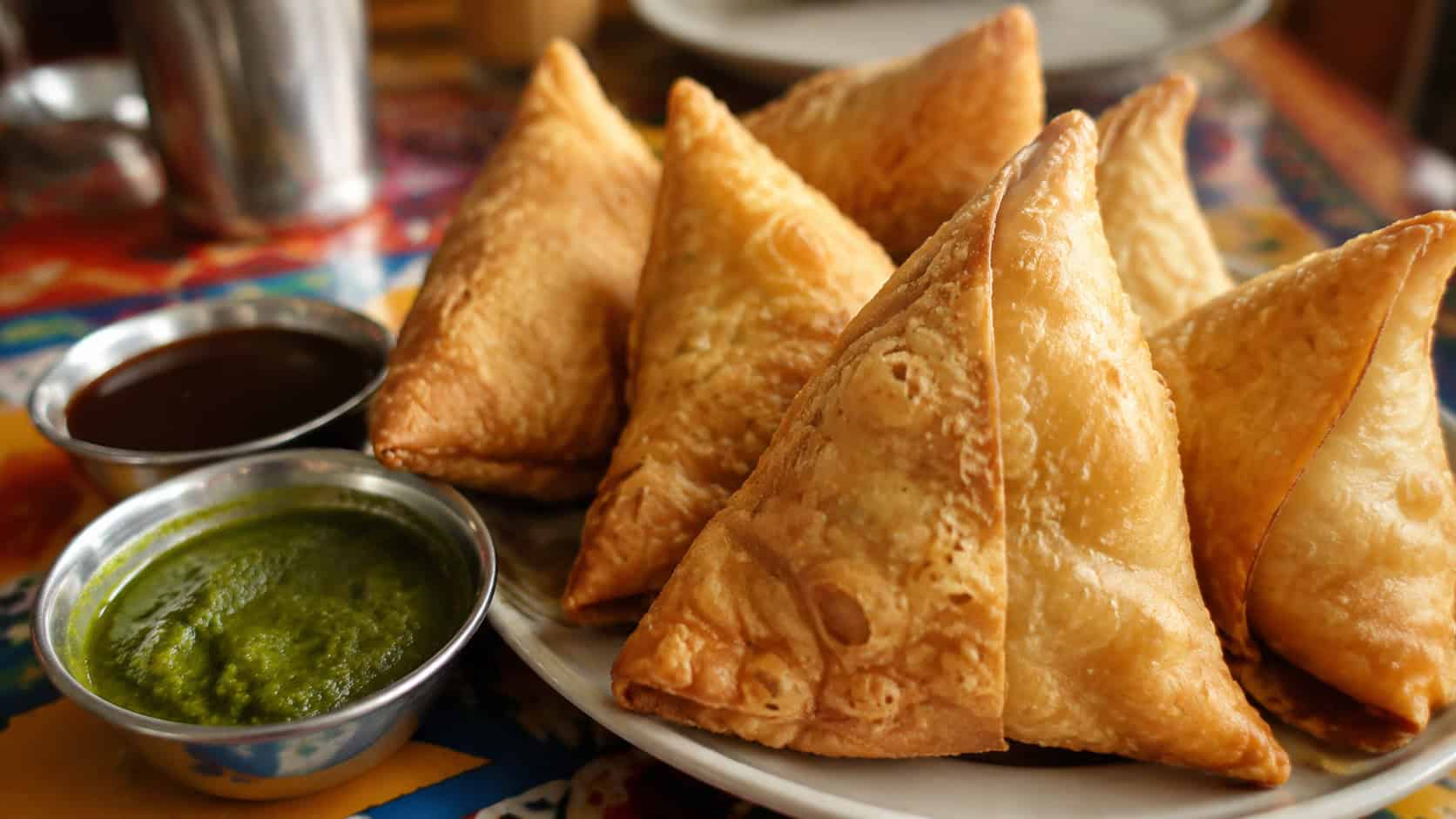 crispy golden samosas with flaky crusts in a wide horizontal shot, piled on a plate with green mint chutney and tamarind sauce on a colorful table, warm natural light, shallow depth of field, landscape orientation