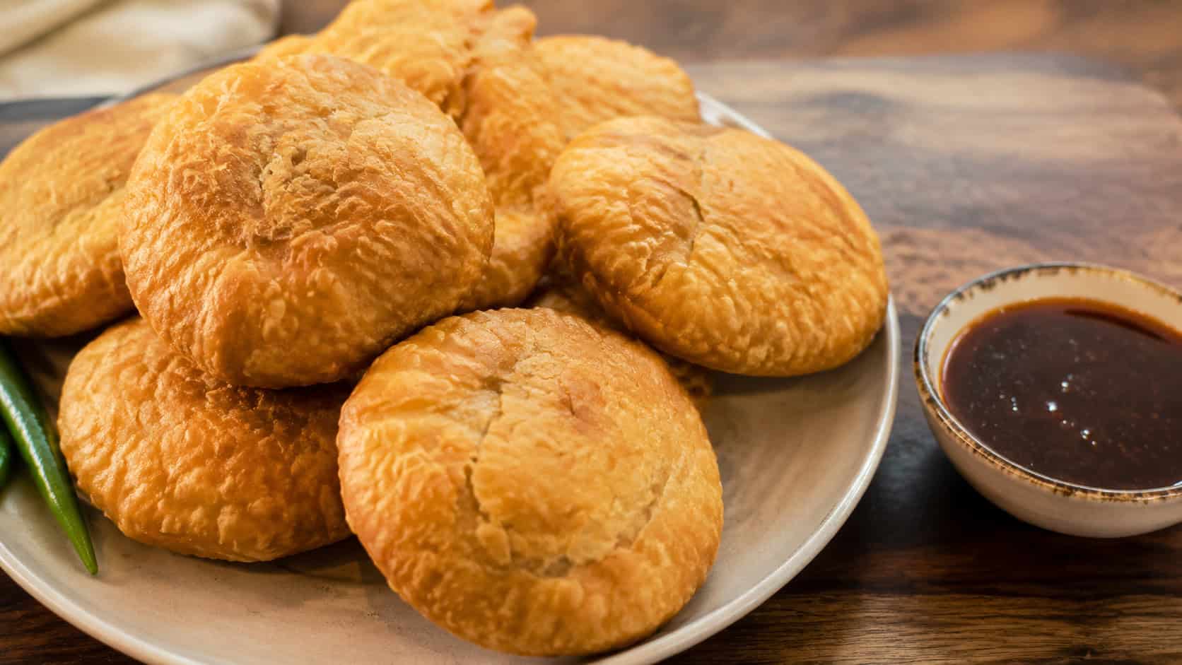 golden flaky kachori pastries in a wide horizontal shot, piled on a plate with tamarind chutney on a wooden board, warm natural light, shallow depth of field, landscape orientation